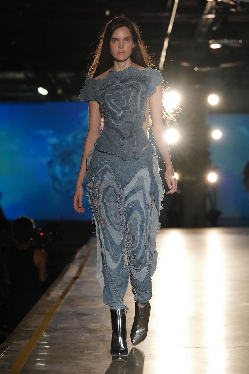Off-shoulder pale denim top, contour-seamed and built from frayed concentric panels. Slouched denim trousers layered with raw-edged blue swirls, a torn opening at one thigh. Black leather ankle boots with mirror-gloss pointed toes. Crossing a floodlit loading dock after tearing down a billboard.