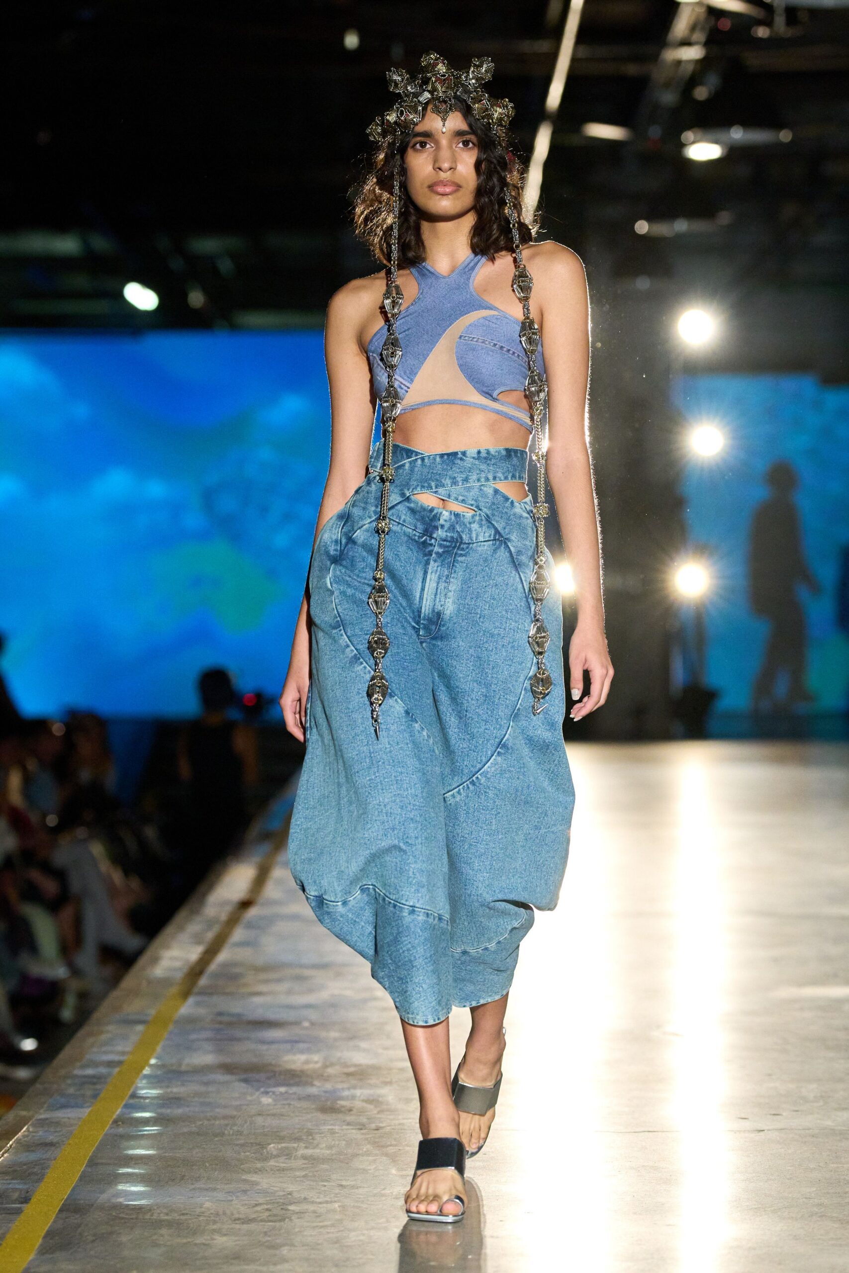 Silver filigree crown with long faceted drops grazing the hips. Powder-blue cutout top in washed denim jersey with nude mesh swoop. Acid-wash denim wrap skirt, waistband crossing over a slashed opening. Flat thong sandals with broad pewter straps. Walking the aquarium tunnel after closing, lights still humming overhead.