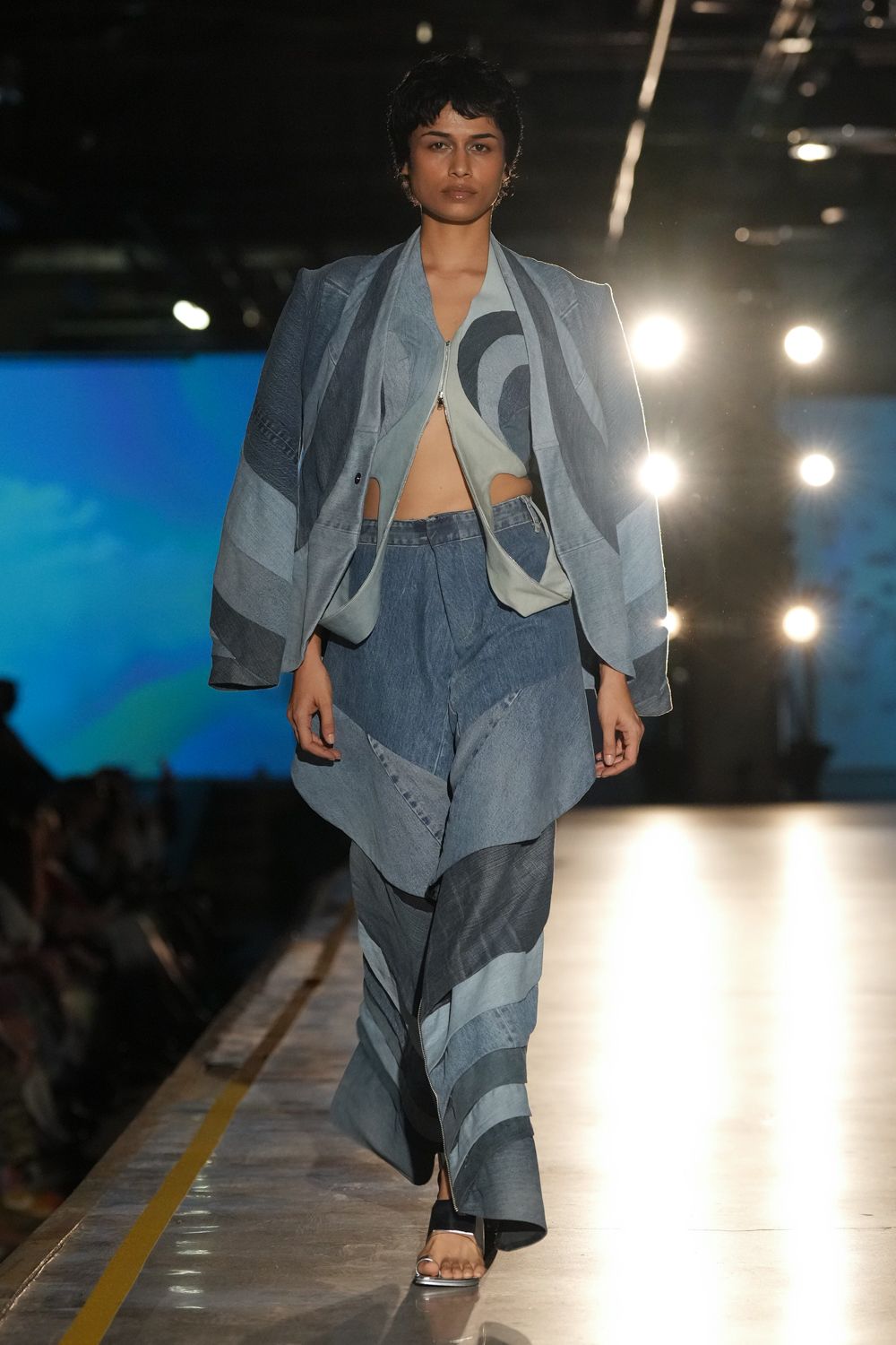 Patchworked indigo jacket in mismatched chambray bands, worn slipping off the shoulders. Pale blue zip-front top cut away at both hips. Wide patchwork denim trousers with a skewed flap panel over the hips. Black toe-loop sandals with a slim white front edge. Crossing an empty ferry deck just after the sun drops.