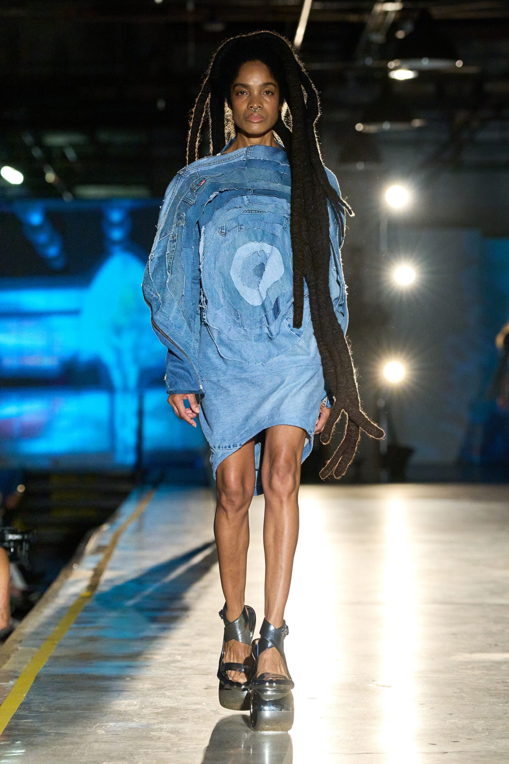 Asymmetric pale denim dress pieced from concentric jacket panels, one seam split raw. Black crisscross platform sandals with thick rounded soles. Walking the loading dock after rain, forklift lights still glaring.