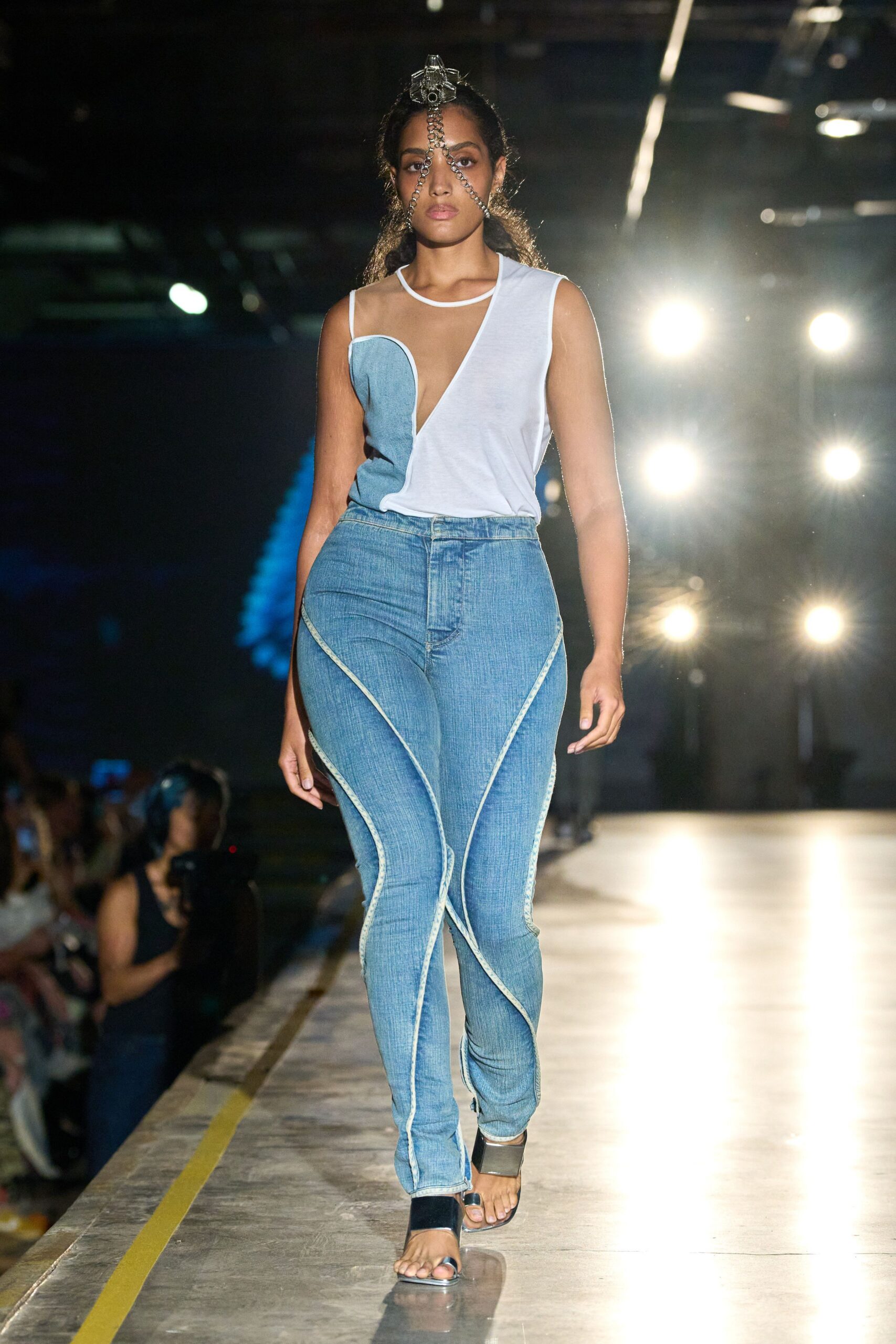 Clear acrylic crest with three chain strands veiling the face. Asymmetric white jersey top sliced by a pale denim bust panel. Bleached skinny jeans with raised curving seams looping down each leg. Black sandals with a broad silver ankle cuff. Walking the long concourse after missing the last train home.