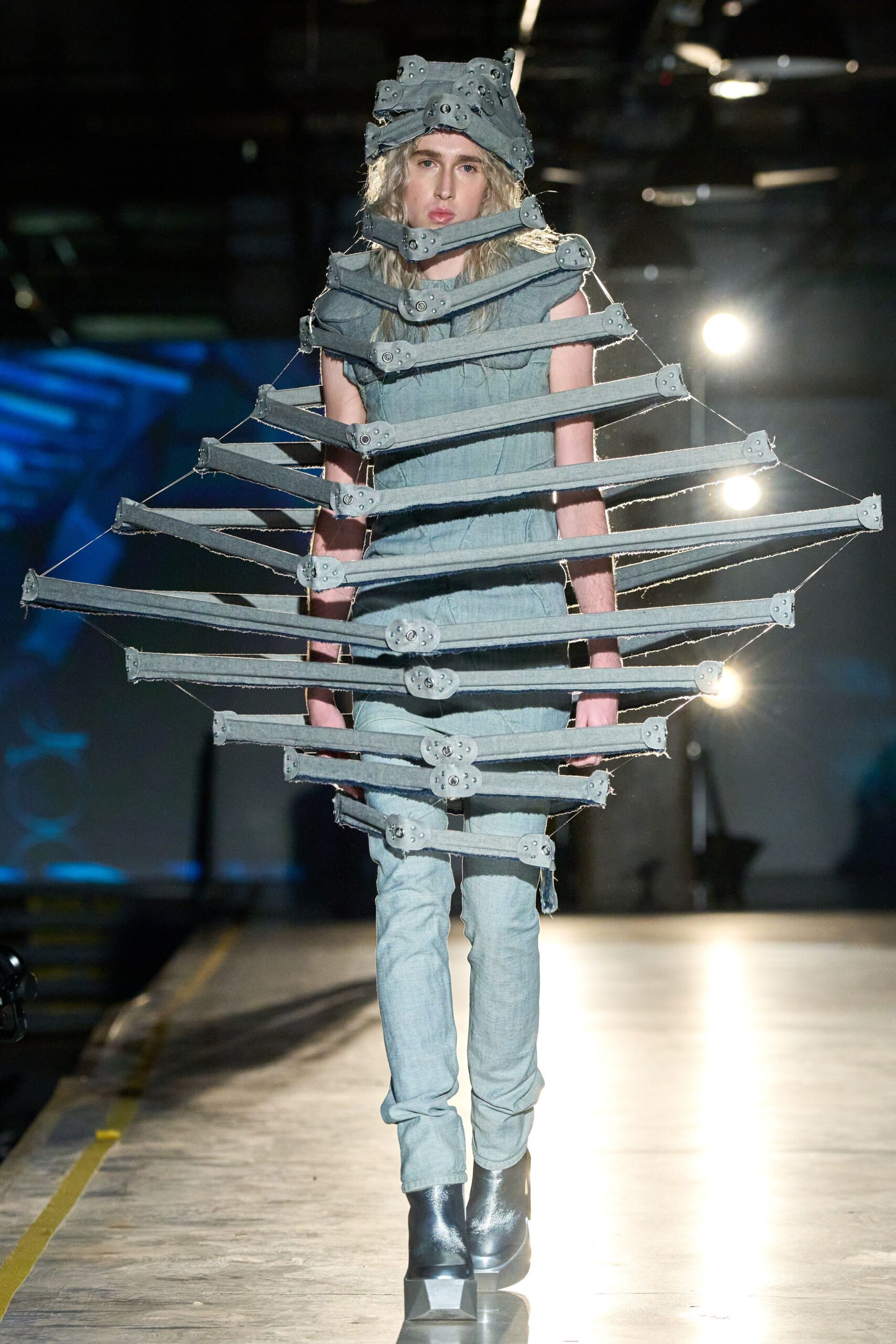 Pale denim bands riveted into a helmet and chin brace. Faded blue denim column under a hoop cage of frayed slats. Bleached skinny jeans, one narrow strip dangling at the calf. Gunmetal platform ankle boots with faceted block toes. Crossing an aircraft hangar after the emergency drill went strange.