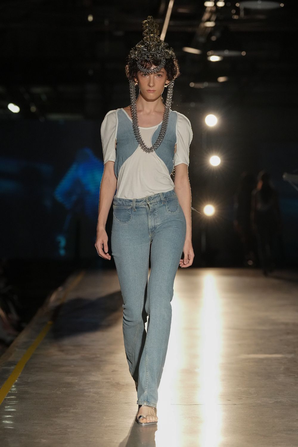 Wirework crown tangled with silver chain, two long loops framing the chest. White jersey top draped off-center over pale denim side panels. Bleached skinny jeans with rounded front pocket stitching. Clear strap sandals, nearly disappearing against the floor. Crossing an empty convention hall after the trade booths shut down.