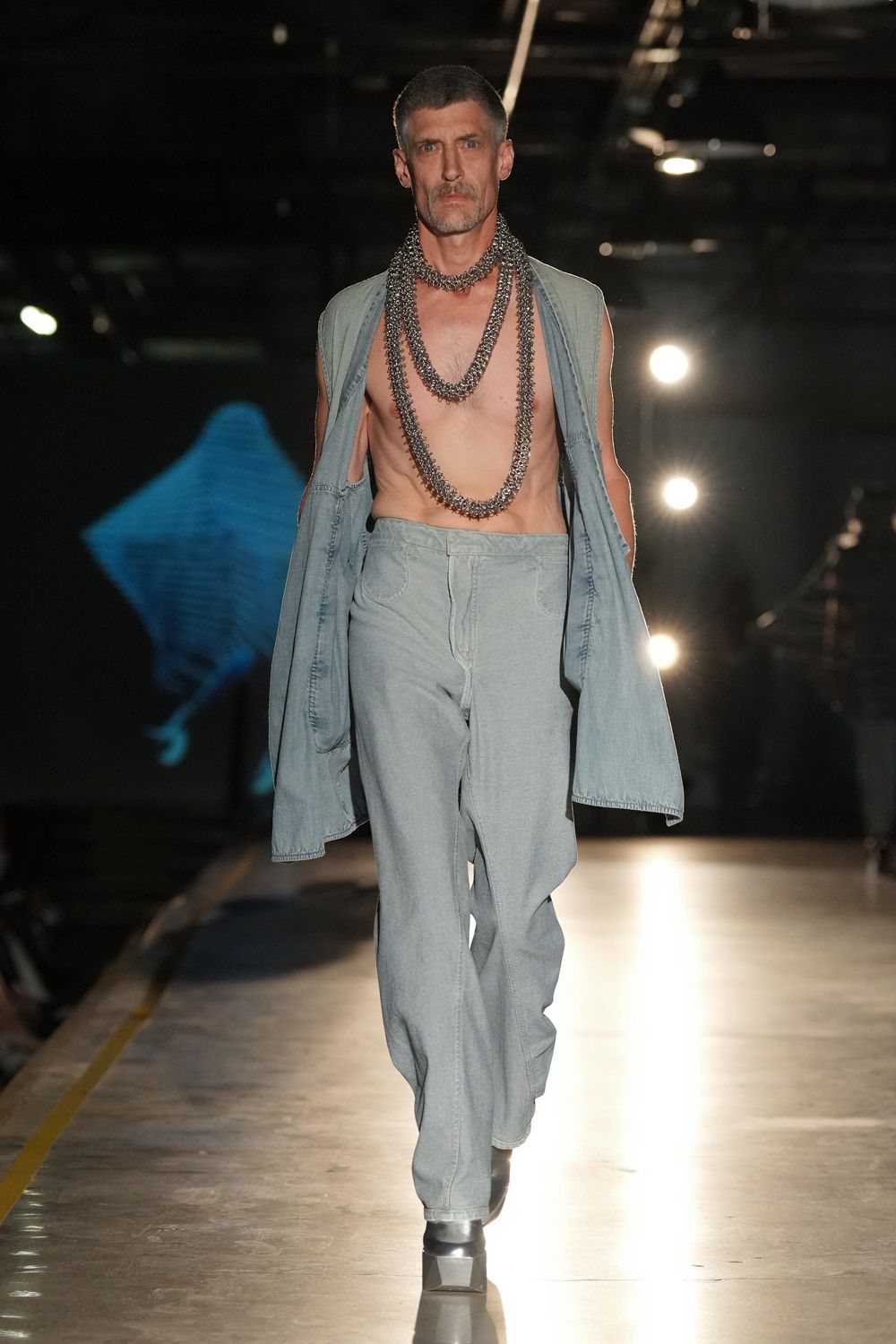 Layered gunmetal curb chains hanging to the navel in three heavy loops. Bleached denim shell left open, armholes cut almost to the waist. Pale denim jeans with a smooth high rise and pooling hems. Black square-toe boots with a clear lucite block heel. Crossing an exhibition hall floor while the rigging lights are still warm.