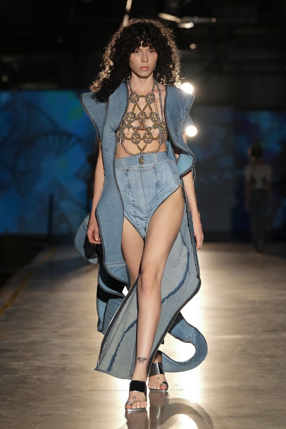 Antique-gold chain harness top, jeweled rosettes linked across the bare chest. Washed denim sleeveless coat with winged shoulders and raw curling edges. High-cut pale denim bodysuit, coin charm hanging at the fly. Black velvet strap mules. Crossing the fairground at dusk after riding something too fast.