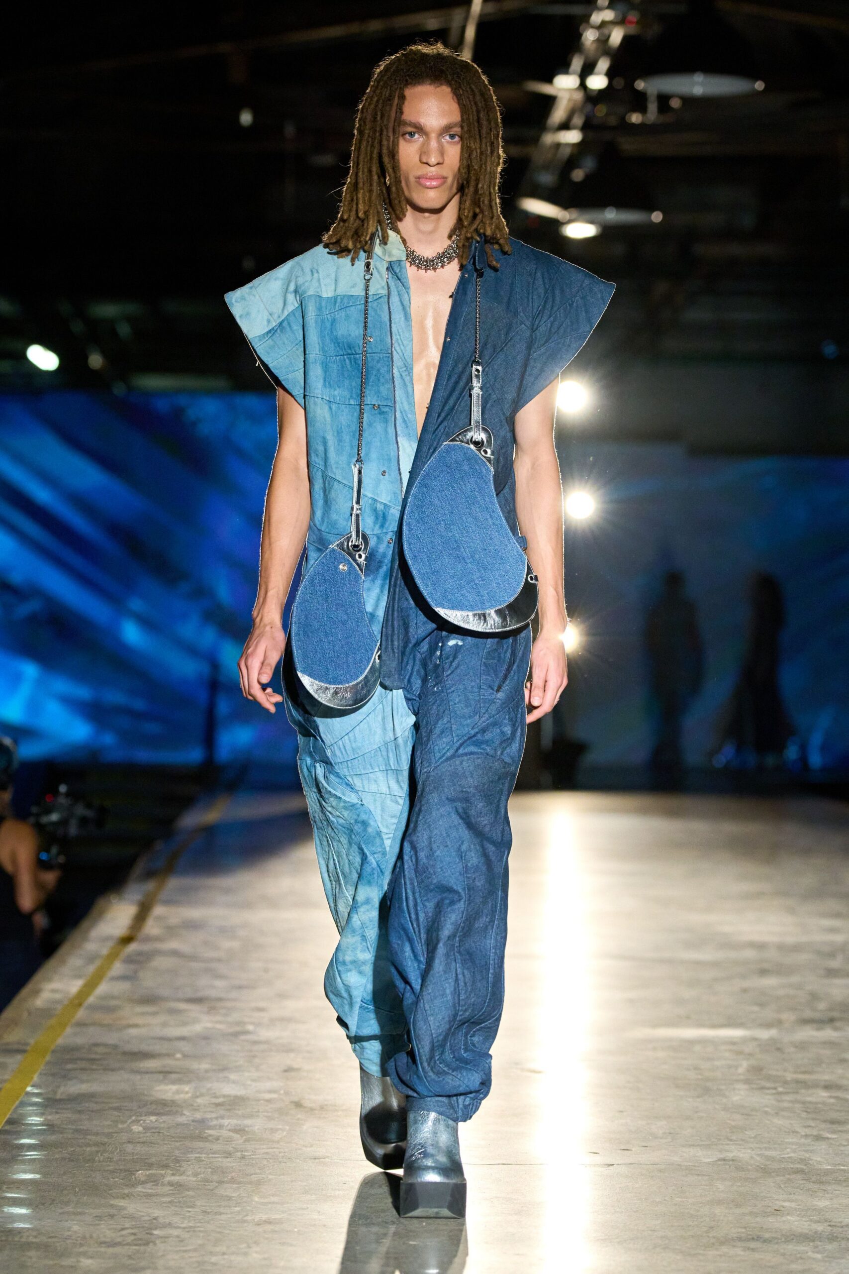 Chunky silver chain sitting tight at the collarbone. Two-tone indigo chambray jumpsuit, raw cap sleeves and a plunging split front. Twin teardrop denim bags hanging low on chain straps. Black platform ankle boots with brushed silver vamps. Crossing the ferry deck after rain, engines still thudding below.