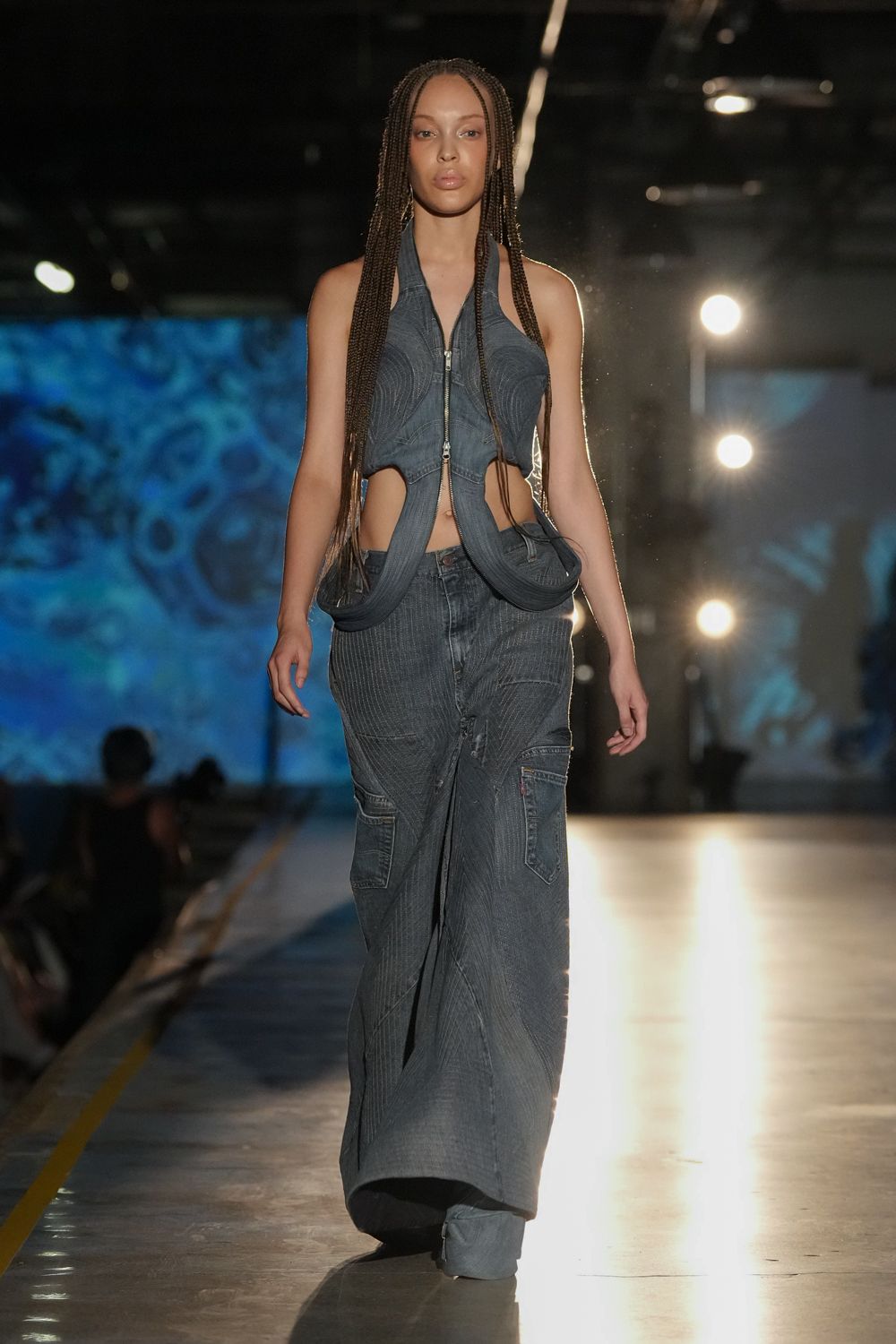 Blue-grey halter top in pinstriped denim, zipped open and looped into hanging bands. Wide pinstriped jeans with curved seam panels and low cargo pockets. Crossing an empty bus depot after rain, headlights still glaring off the concrete.