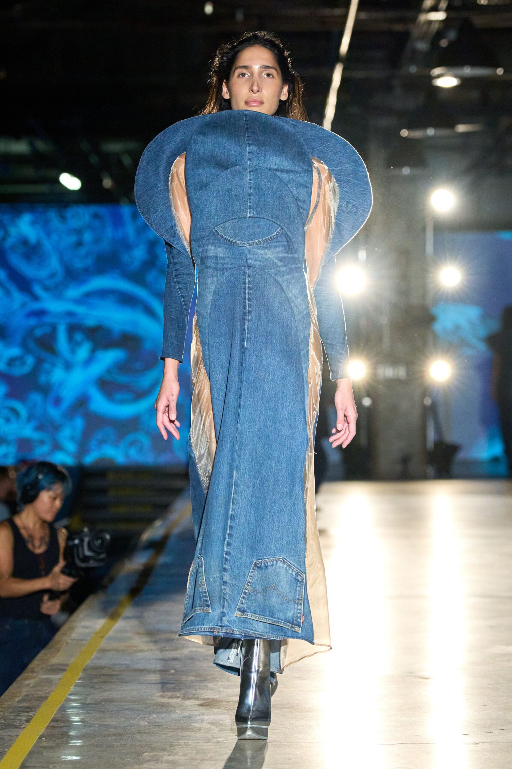 Sculptural washed-denim column dress with disc-like shoulder flares and jean-pocket appliqué. Champagne sheer organza panels glinting along the sides. Black leather ankle boots with mirrored wedge soles. Walking under strip lights in a disused depot after the floor has been hosed down.