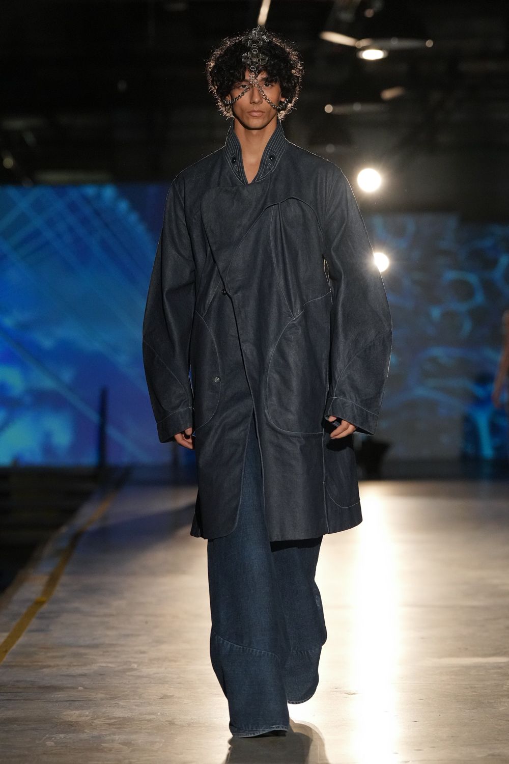 Black chain face harness climbing from brow to jaw in linked loops. Ink denim coat with curved panel seams and a doubled stand collar. Dark indigo wide-legs breaking over the shoe with a deep hem panel. Crossing an aircraft hangar after midnight, engines still ticking cool.