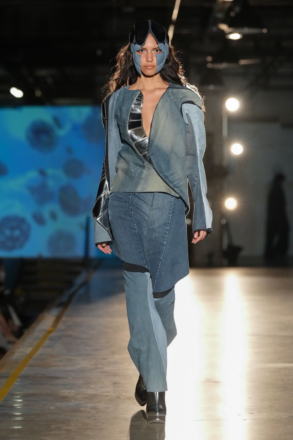 Faded denim face guard topped with black goggle-shaped panels. Asymmetric washed-denim jacket, plunging open and edged with black leather strips. Patchworked blue denim trousers with curved black bands at the knees. Black square-toe ankle boots. Crossing an aircraft hangar floor after everyone else has gone.