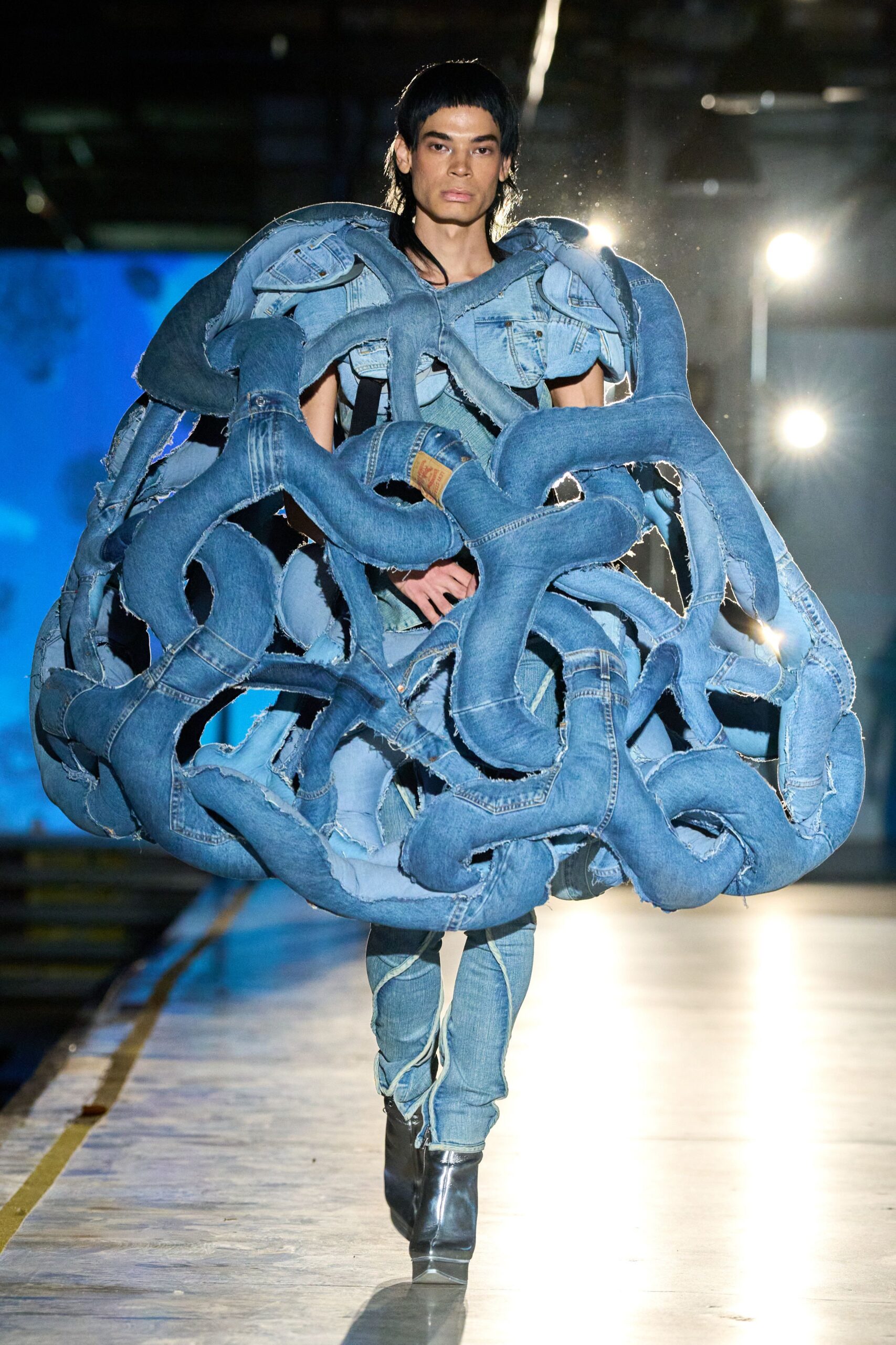 Bulbous cage of stuffed blue jeans, frayed seams and a tan back patch. Pale denim leggings with spiral seam lines wrapping the calves. Black leather ankle boots with square toes. Crossing a loading dock after rain, carrying the whole scrap heap with you.