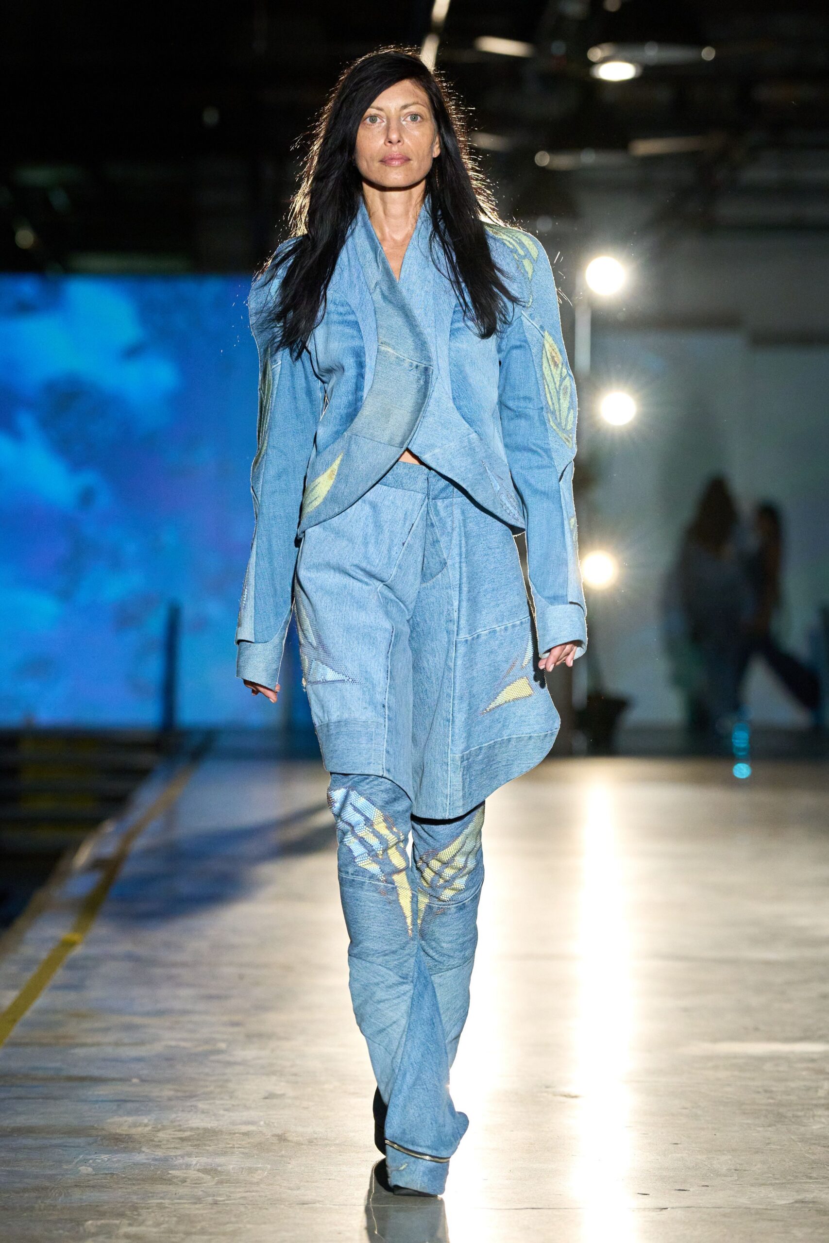 Faded blue denim jacket with a draped shawl front and raw pieced seams. Matching denim trousers, curved paneling and pale yellow intarsia at the knees. Pointed shoes swallowed under extra-long flared hems. Crossing an aircraft hangar at dawn while the floodlights are still on.