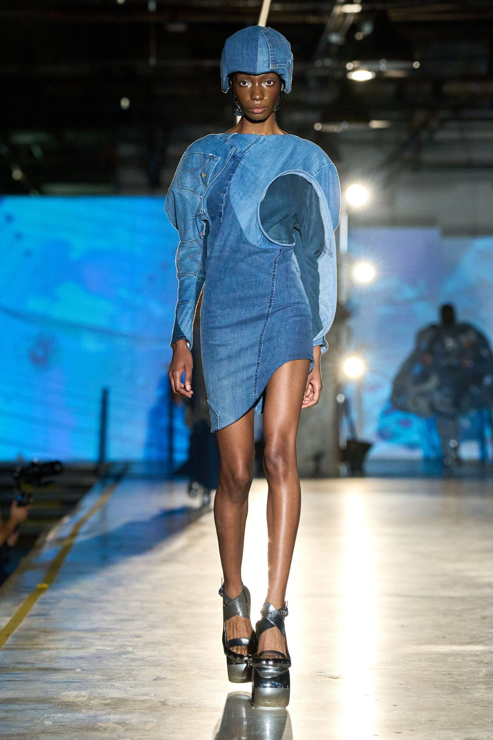 Padded denim aviator cap with ear flaps hanging loose. Spliced blue denim mini, one shoulder carved into a giant armhole. Black patent platforms with crossed straps and a chrome-bright sole. Underpass after rain, headlights catching the chalk seams.