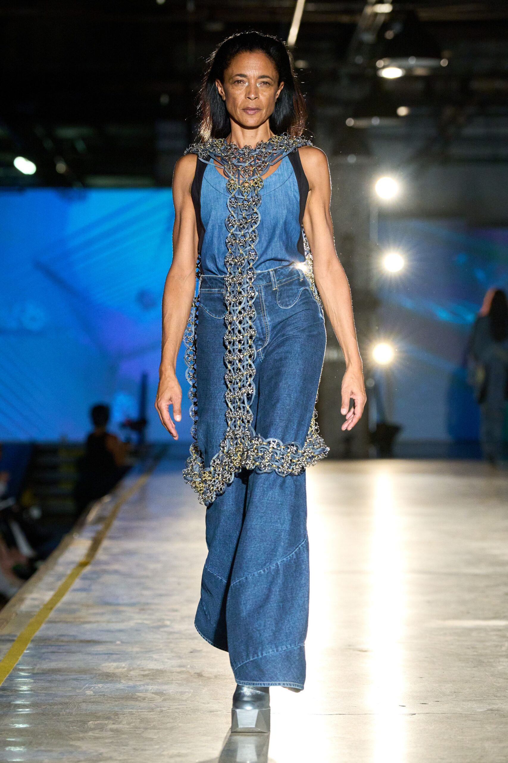 Cascading lattice bib of linked metal rosettes draped to the knee. Faded indigo chambray tank, cut low under the arms. Dark denim wide-legs with curved seaming and a high waist. Square black boots with blunt blocky toes. Crossing a ferry deck after dusk, salt drying on your shoulders.
