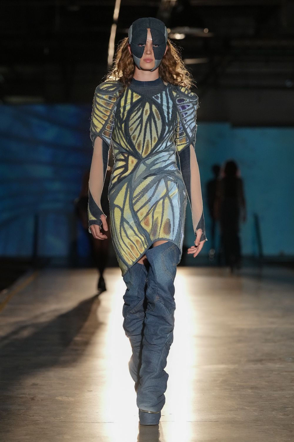 Dark padded face mask, split open at one eye and mouth. Butterfly-wing knit dress with ruched seams and cutout side panels. Matching knit arm warmers, thumbholes stretched and fraying. Denim thigh-high boots, heavily slouched from shin to ankle. Crossing the aquarium tunnel after the school trip has emptied out.