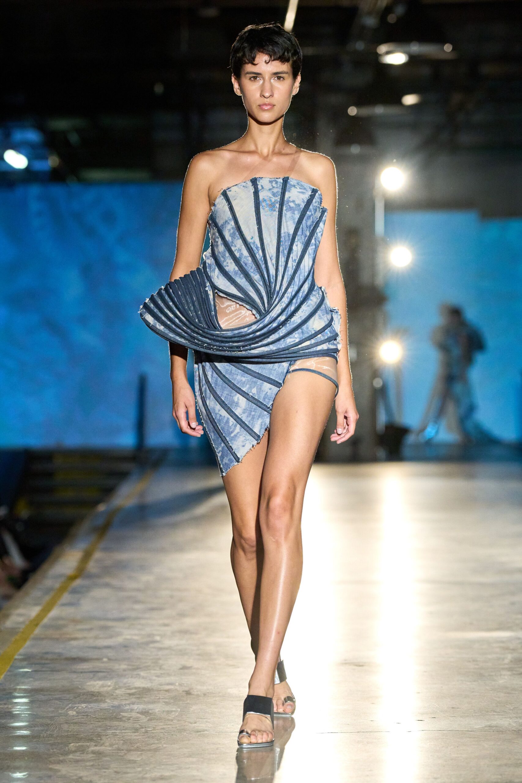 Strapless cloud-wash mini with navy boning channels spiraling into a sculpted hip fan. Sheer nude briefs, one side caught by a single blue strap. Black toe-loop sandals with mirrored block heels. Crossing an empty pool deck after the storm blew through.