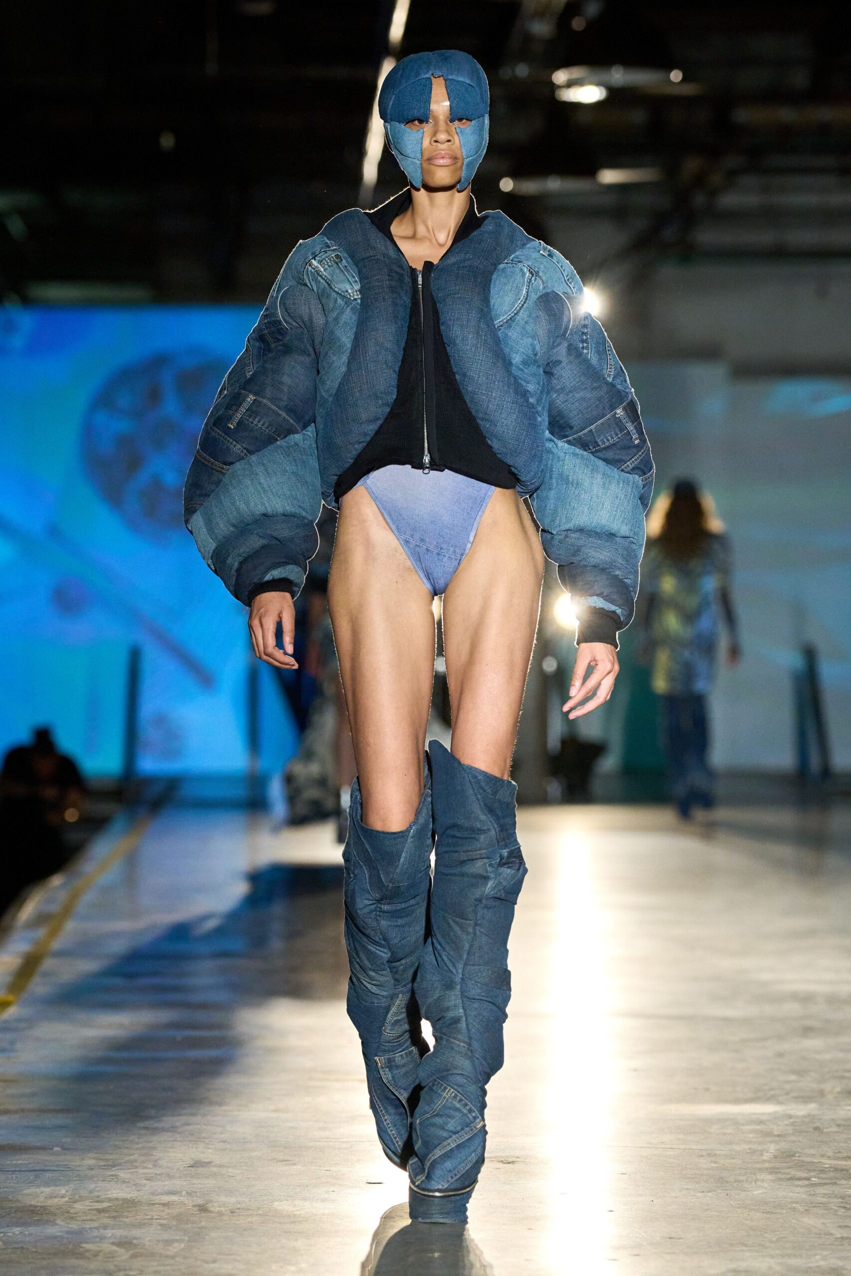 Padded indigo denim helmet, split open at the forehead and eyes. Bulbous patchwork denim bomber, pieced from faded jean panels. Pale blue briefs cut high on the hip. Denim thigh-high boots, slouched and spiraled with jean seams. Crossing an empty ice rink after the generators finally kick in.