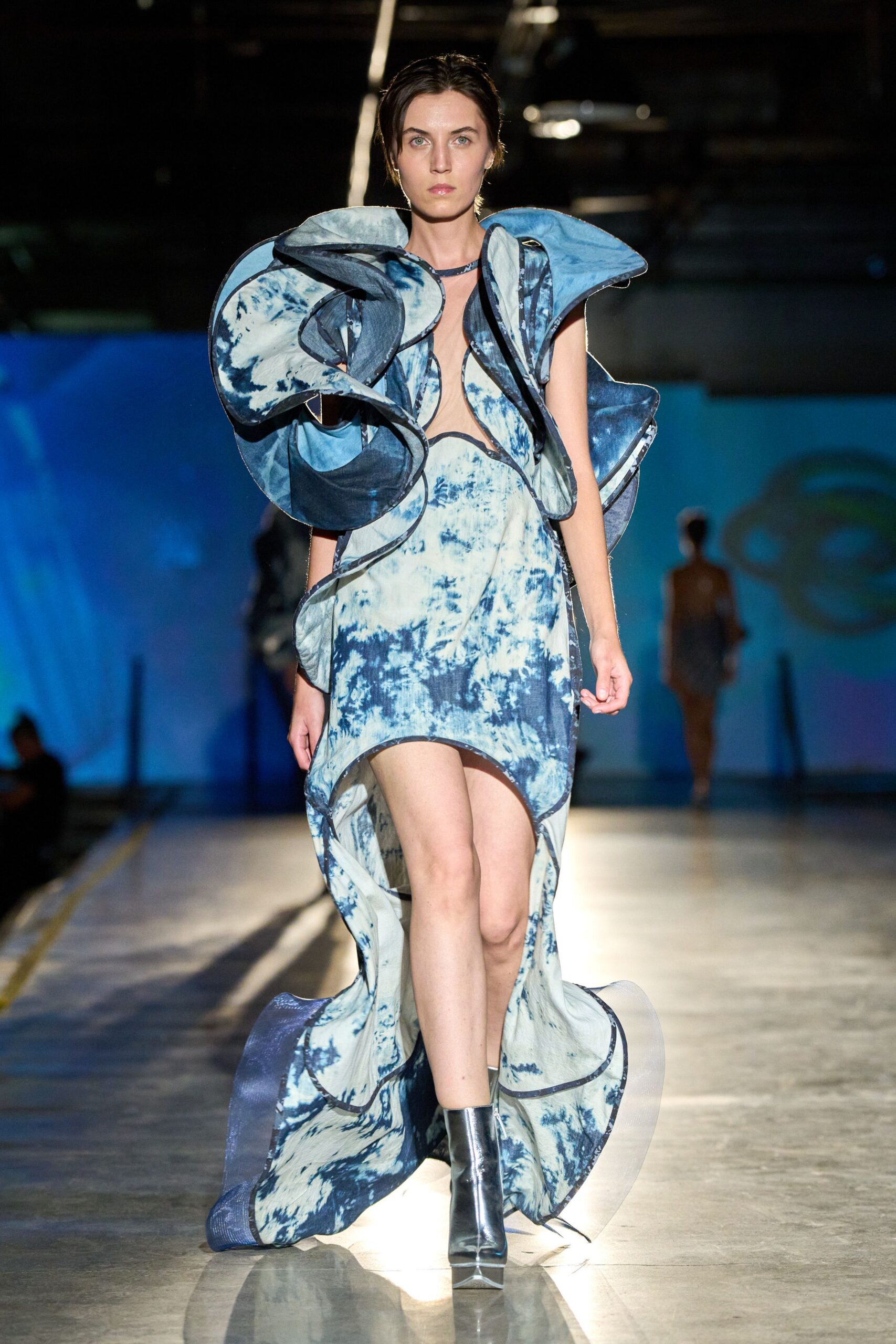 Indigo tie-dye gown exploding into wired organza ruffles around the shoulders. Mirror-silver ankle boots with a blunt square toe. Storm exhibit at the science museum, school groups just cleared out.