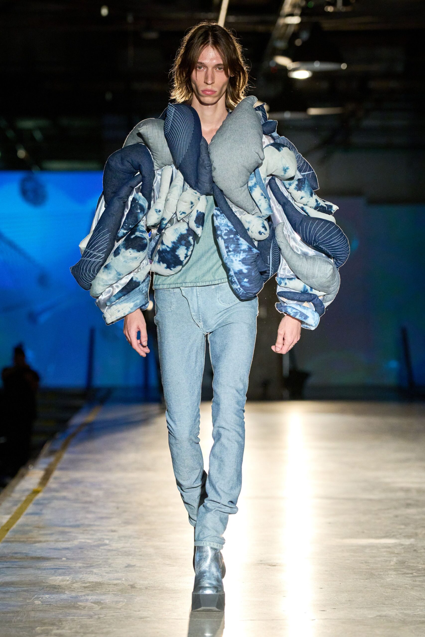 Oversized jacket swarmed with stuffed tubular pads in indigo shibori and pinstripe. Bleached skinny jeans, whiskers ghosting across the high thigh. Square-toe silver boots with a heavy flared platform. Crossing an empty loading dock wrapped in borrowed bedding.