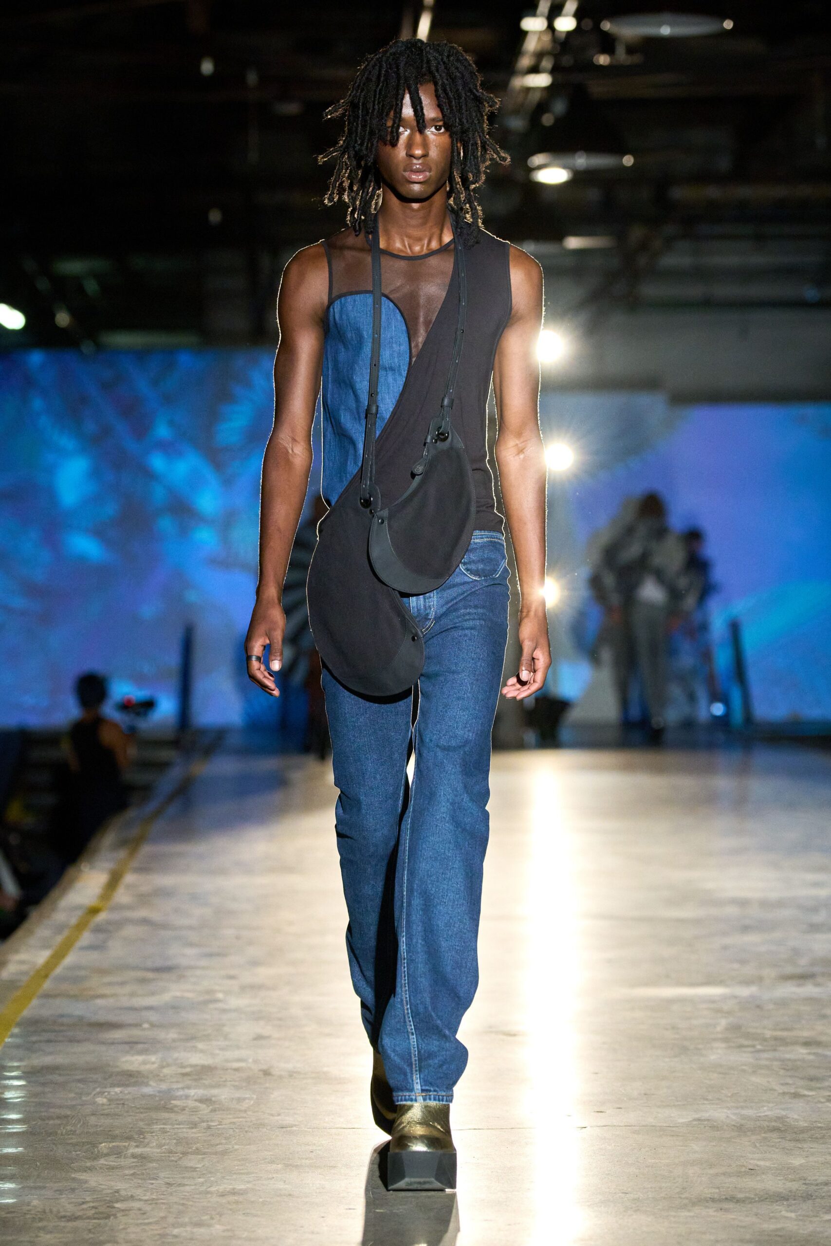 Asymmetric black jersey top with a sheer mesh yoke and denim inset. Two black crescent pouches hanging low on thin buckled straps. Mid-wash jeans with a curved pocket peeking under the draped hem. Square-toe black boots with chunky wedge fronts. Walking out of a warehouse set while the work lights are still up.