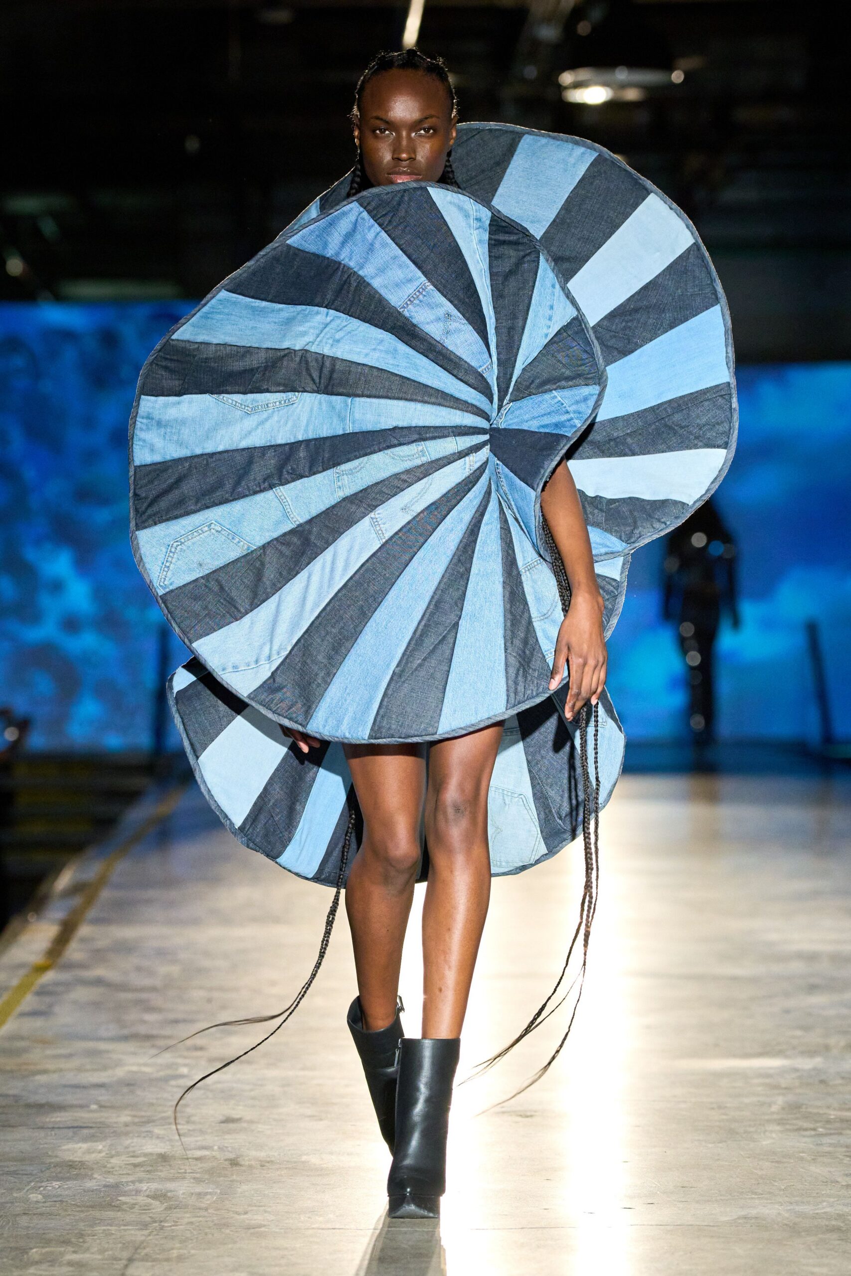 Spiral cocoon dress pieced from pale and dark denim wedges, pocket tops still visible. Black ankle boots with hard square toes. Crossing an aircraft hangar with propellers still winding down.