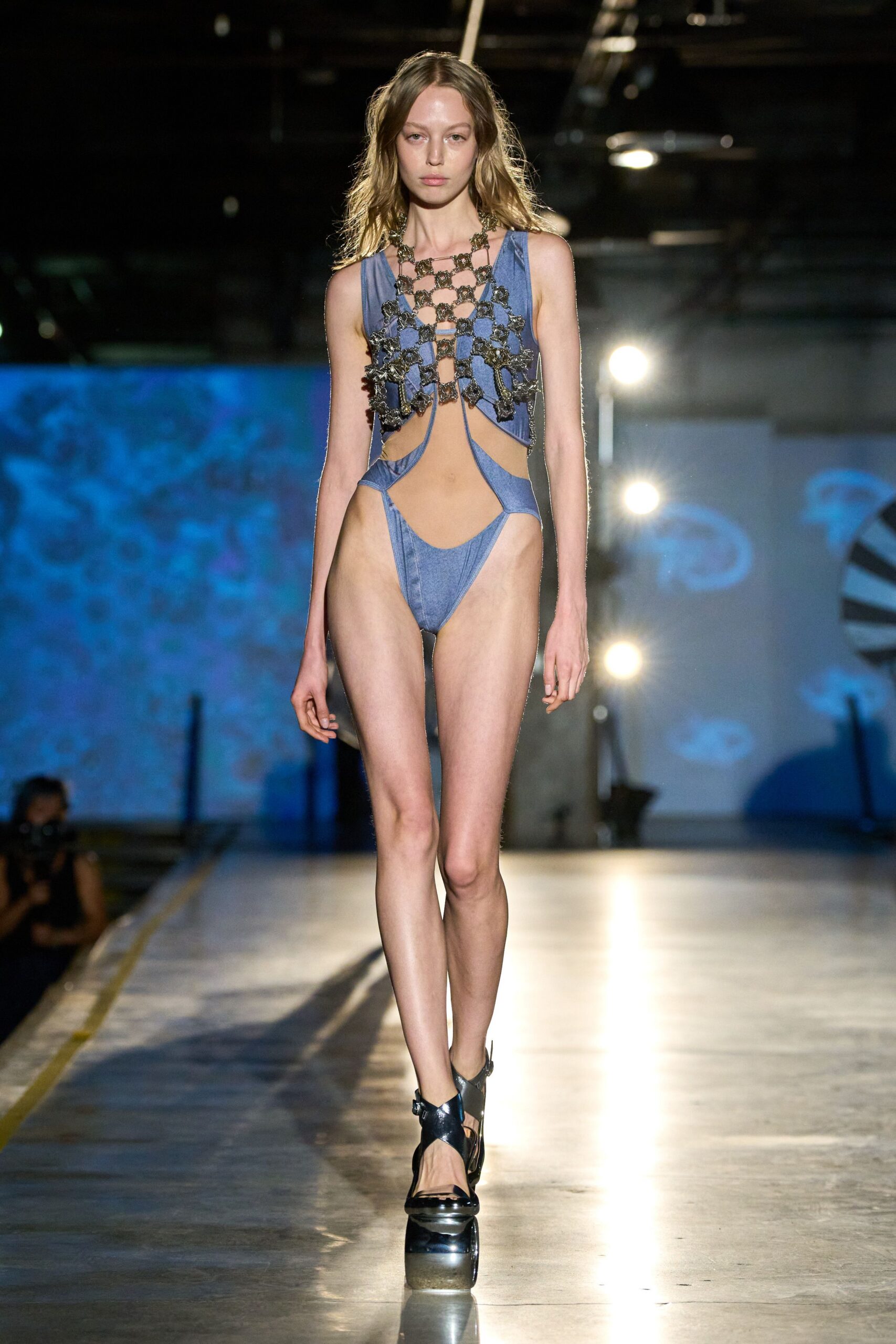 Oxidized metal lattice bib with flowered links draped over the chest. Faded blue jersey monokini cut into deep side crescents and a high thong. Black patent slingbacks with a thick mirrored platform. Pool deck after midnight, concrete still warm under bare shoulders.