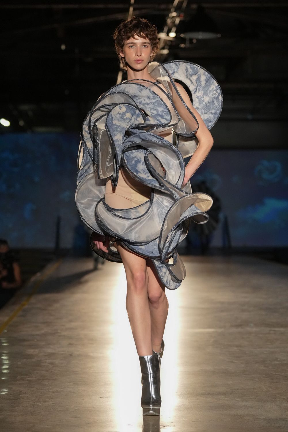 Sheer organza spirals cloud-printed in blue grey, each edge bound dark. Nude base layer with one thin black strap crossing the chest. Mirror-silver ankle boots with a hard square toe. Under the planetarium projector after the school trip has gone home.