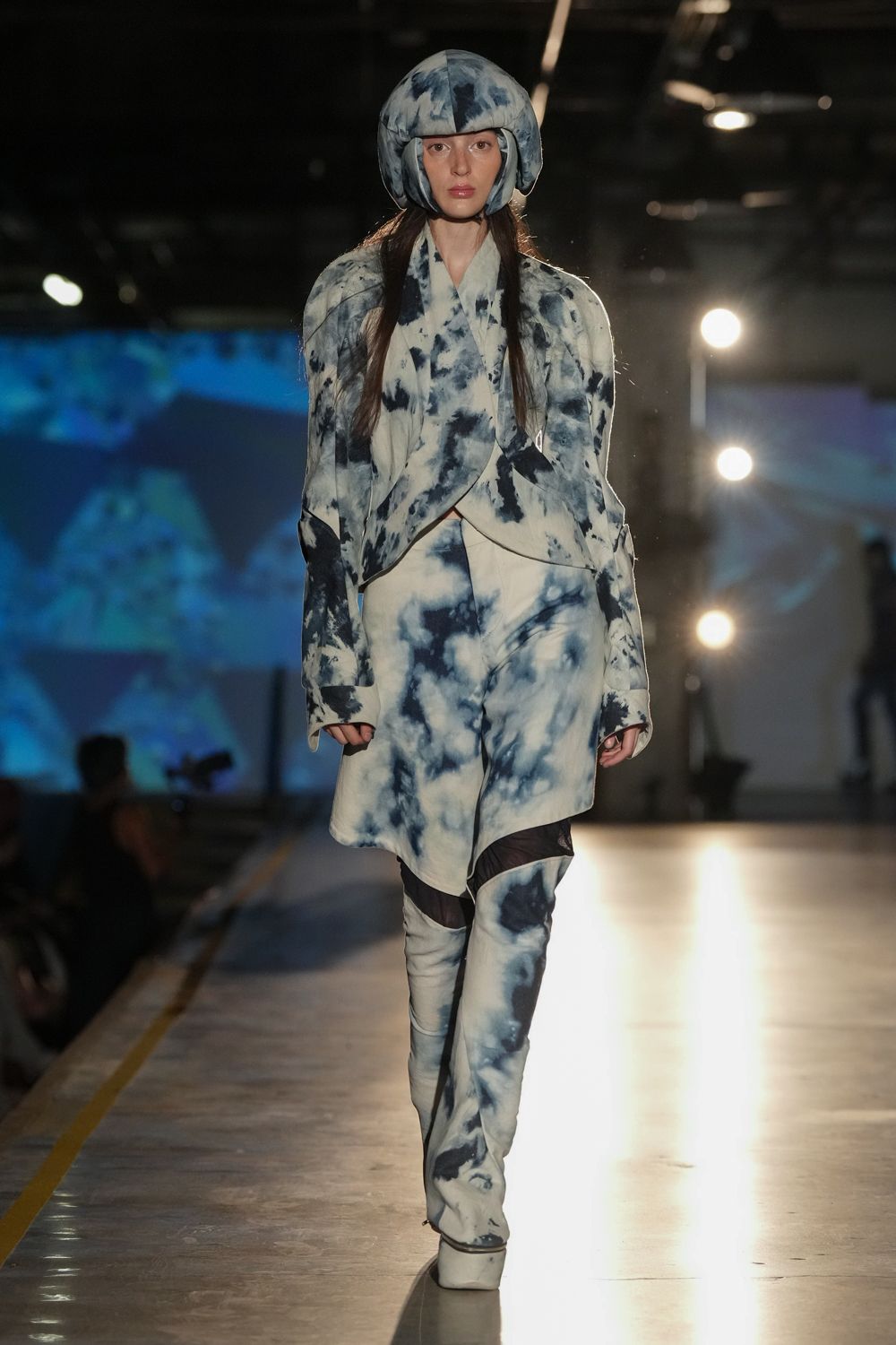 Padded dome helmet in cloudy indigo tie-dye. Cream wrap jacket washed with inky shibori, hem crossing low. Asymmetric skirt panel in the same blurred dye pattern. Thigh-high platform boots broken by sheer black mesh at the knee. Crossing the ferry deck before sunrise, salt stuck to everything.