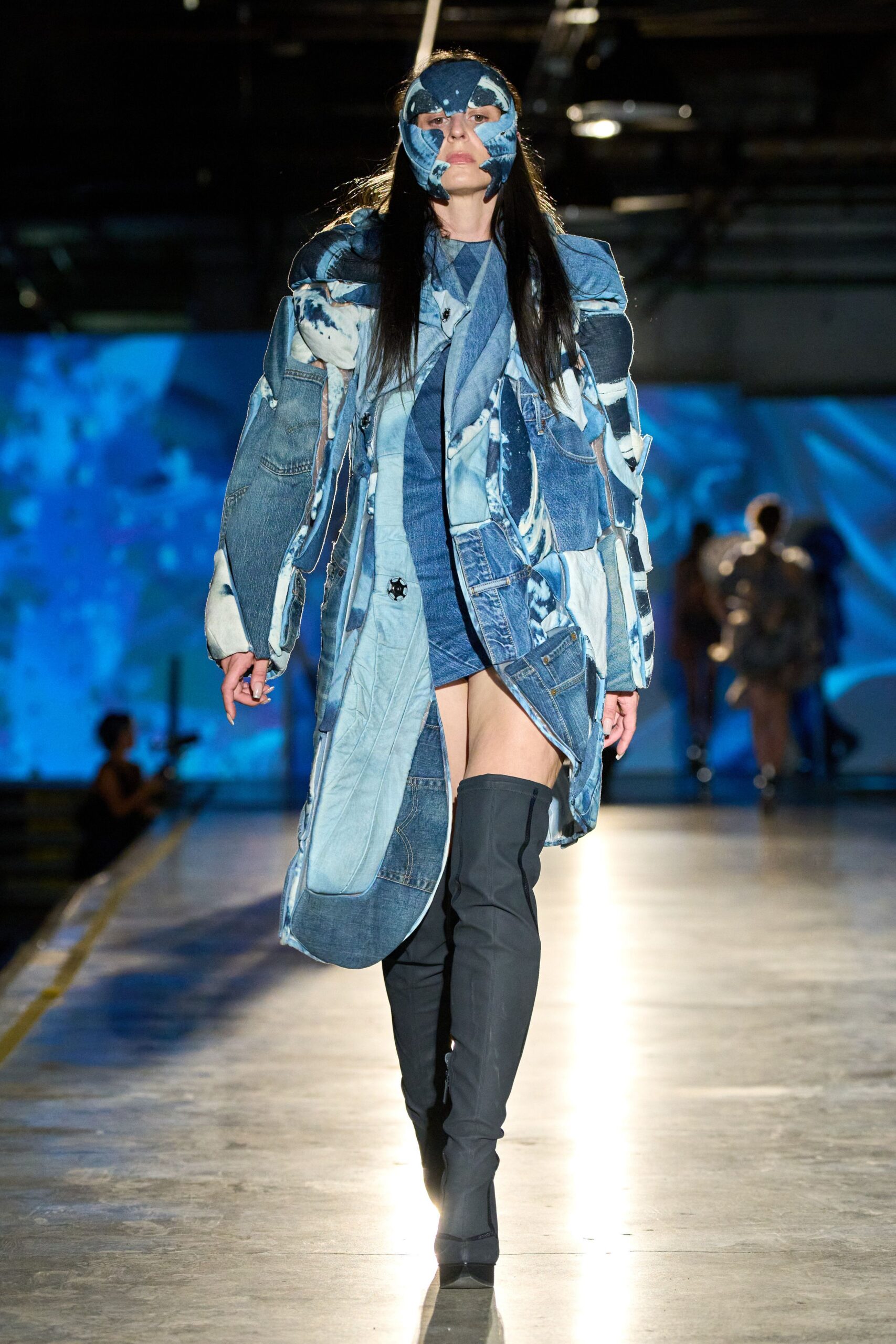 Patchworked blue denim mask, raw-edged around the cheeks and brow. Long coat built from bleached jean panels, pockets and seams colliding. Asymmetric blue denim mini, one side cut high at the thigh. Matte black thigh-high boots with a narrow pointed toe. Crossing the aquarium tunnel after closing, floor still wet from the mop.
