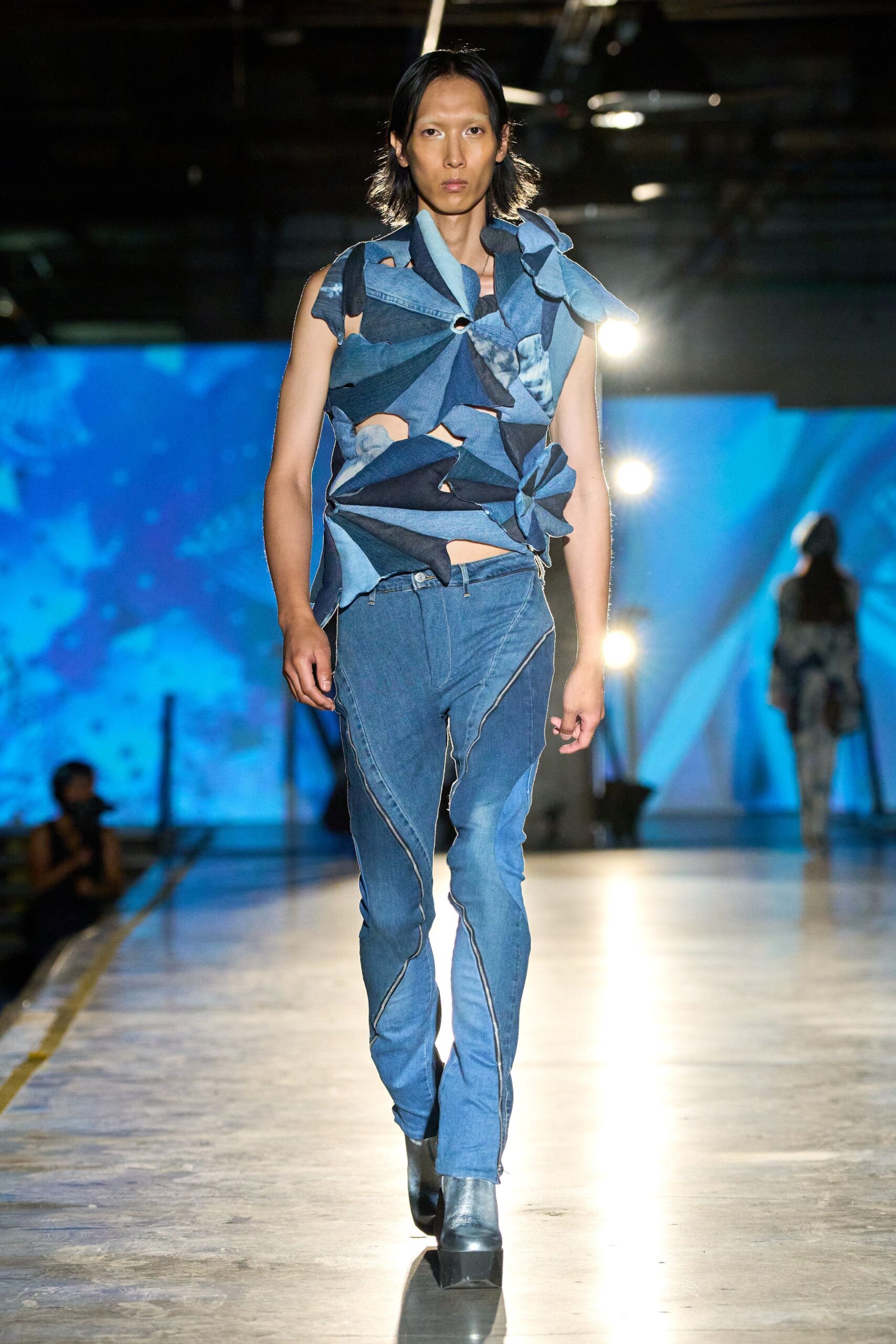 Patchworked indigo denim petals spiraled from a ring, cut open at the ribs. Faded blue jeans traced with curved contrast seams, one hem dragging loose threads. Black square-toe leather ankle boots. Crossing the ferry deck while the wind snaps everything sideways.