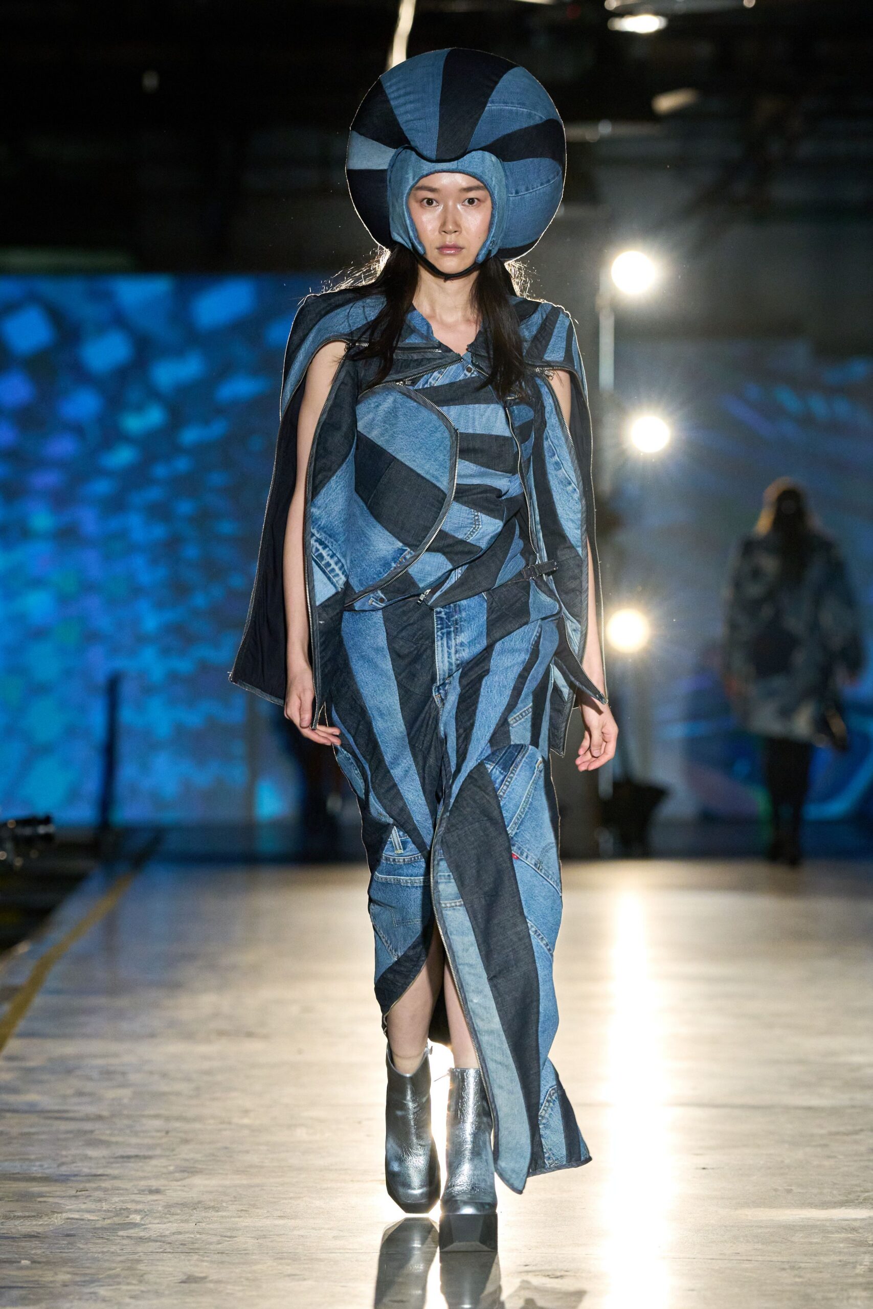 Padded spiral helmet in pieced indigo and washed-blue denim. Asymmetric patchwork denim dress, silver zips tracing the draped seams. Gunmetal ankle boots with blunt square platforms. Crossing a ferry deck after dark, wind flattening the harbor water.