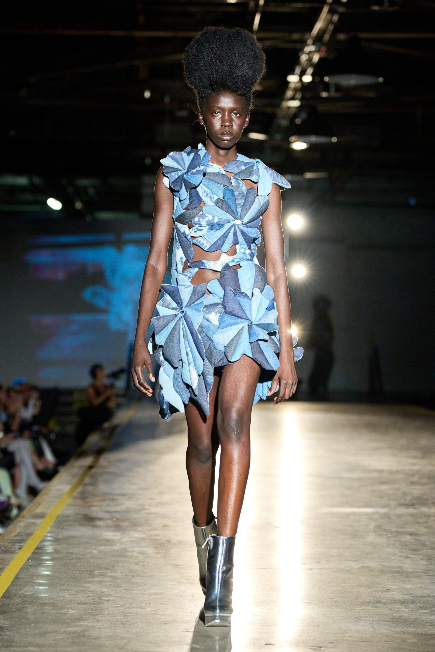 Cutout mini built from pinwheeling blue denim petals, one shoulder ruffled high. Mirror-silver ankle boots with squared toes. Crossing an empty depot floor while welding sparks jump somewhere behind you.