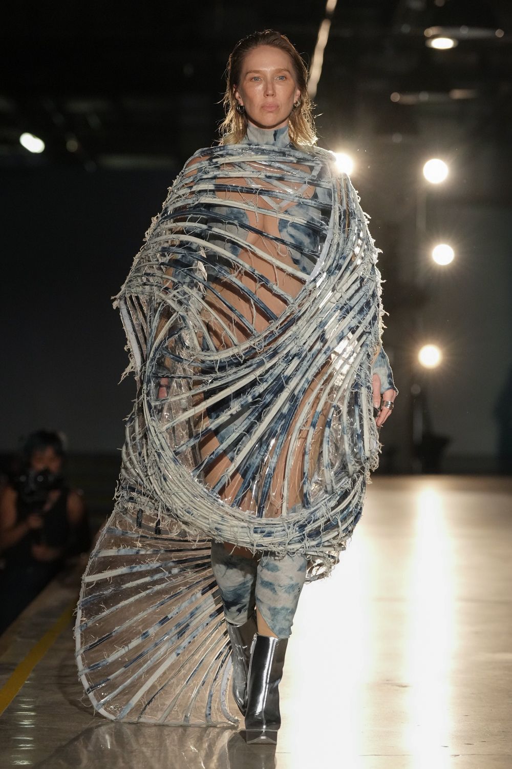Transparent vinyl cocoon ribbed with frayed indigo and chalk denim strips. Sheer nude bodysuit under a smoky tie-dye high neck. Clouded blue-grey leggings with a cutout exposing one knee. Mirror-shine silver boots with blunt square toes. Stacked silver rings. Crossing a ferry deck in wind that turns everything into rigging.