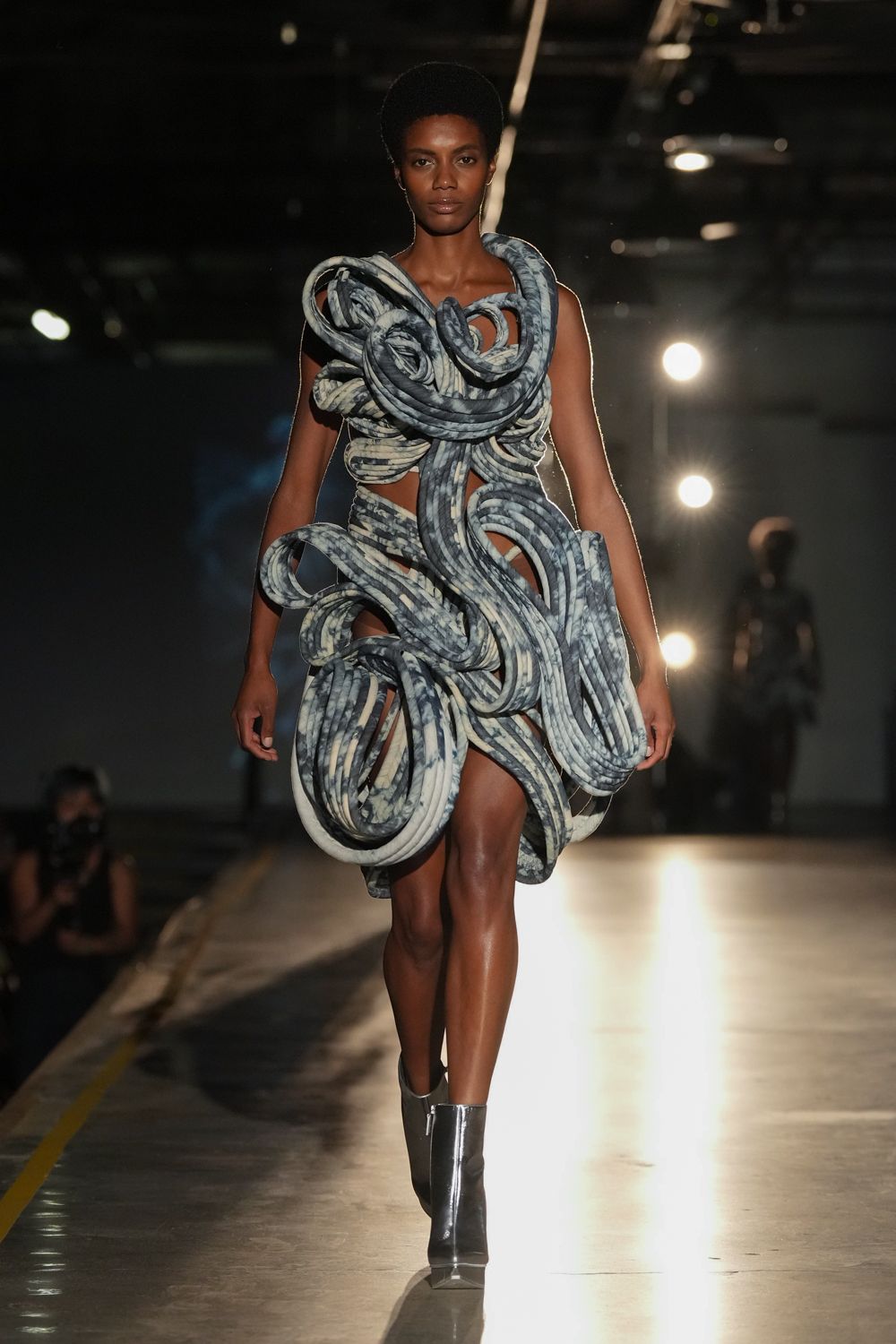 Blue-grey marbled tubular dress knotted into huge looping coils, bare skin between them. Shiny charcoal ankle boots with a narrow white center seam. Crossing a floodlit depot floor after midnight, still carrying all the cables.
