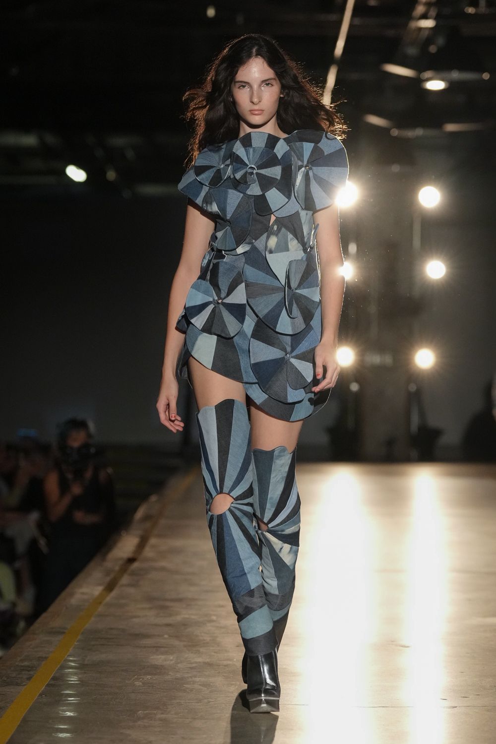 Blue patchwork mini built from pinwheel circles, one shoulder jutting wide. Matching paneled boots with oval knee cutouts and black square toes. Crossing an empty expo hall after the lights have clicked off.