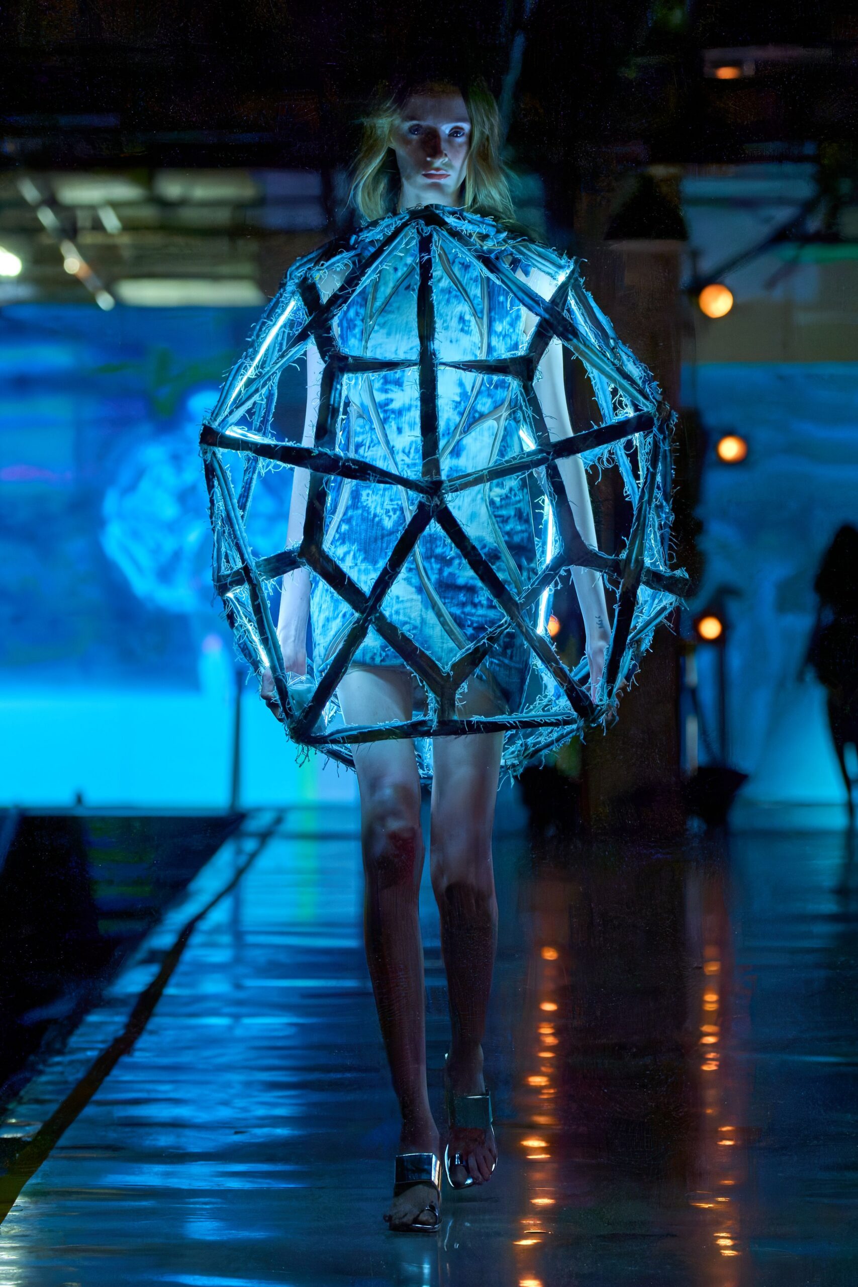 Blue-white iridescent shell trapped inside a black geometric frame, frayed at every edge. Silver mule heels with broad mirrored straps. Crossing a hotel pool deck after midnight rain, lit electric blue.