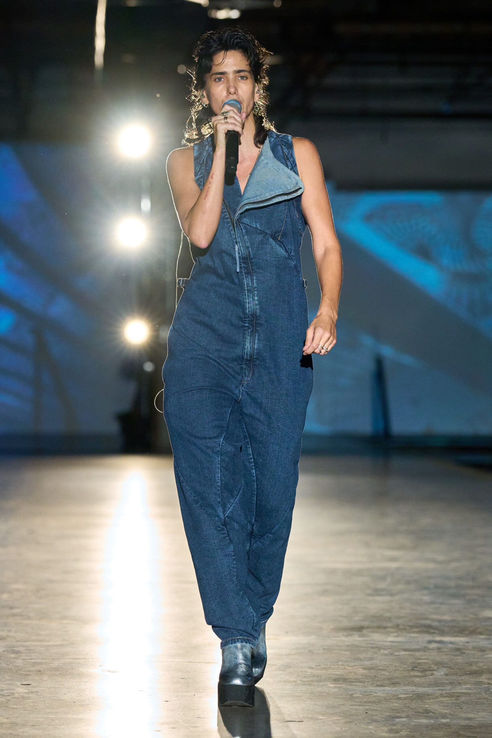 Dark indigo denim jumpsuit with a wide asymmetric lapel and curved seaming. Black wedge ankle boots, blunt and glossy at the toe. Silver ring catching the microphone hand. Soundcheck in an exhibition hall, house lights still glaring.