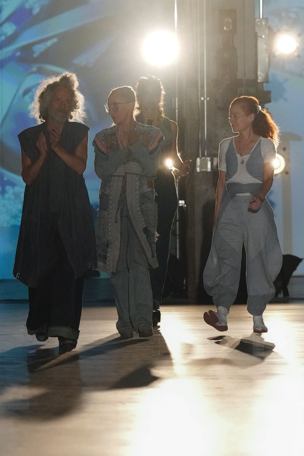 White cap-sleeve tee with pale blue curved inset panels. Powder-blue jumpsuit cut with dropped crotch and gathered cuffed ankles. White sneakers with brick-red soles flashing mid-step. Small silver pendant hanging at the V neck. Crossing a gym floor after the community dance showcase lets out.