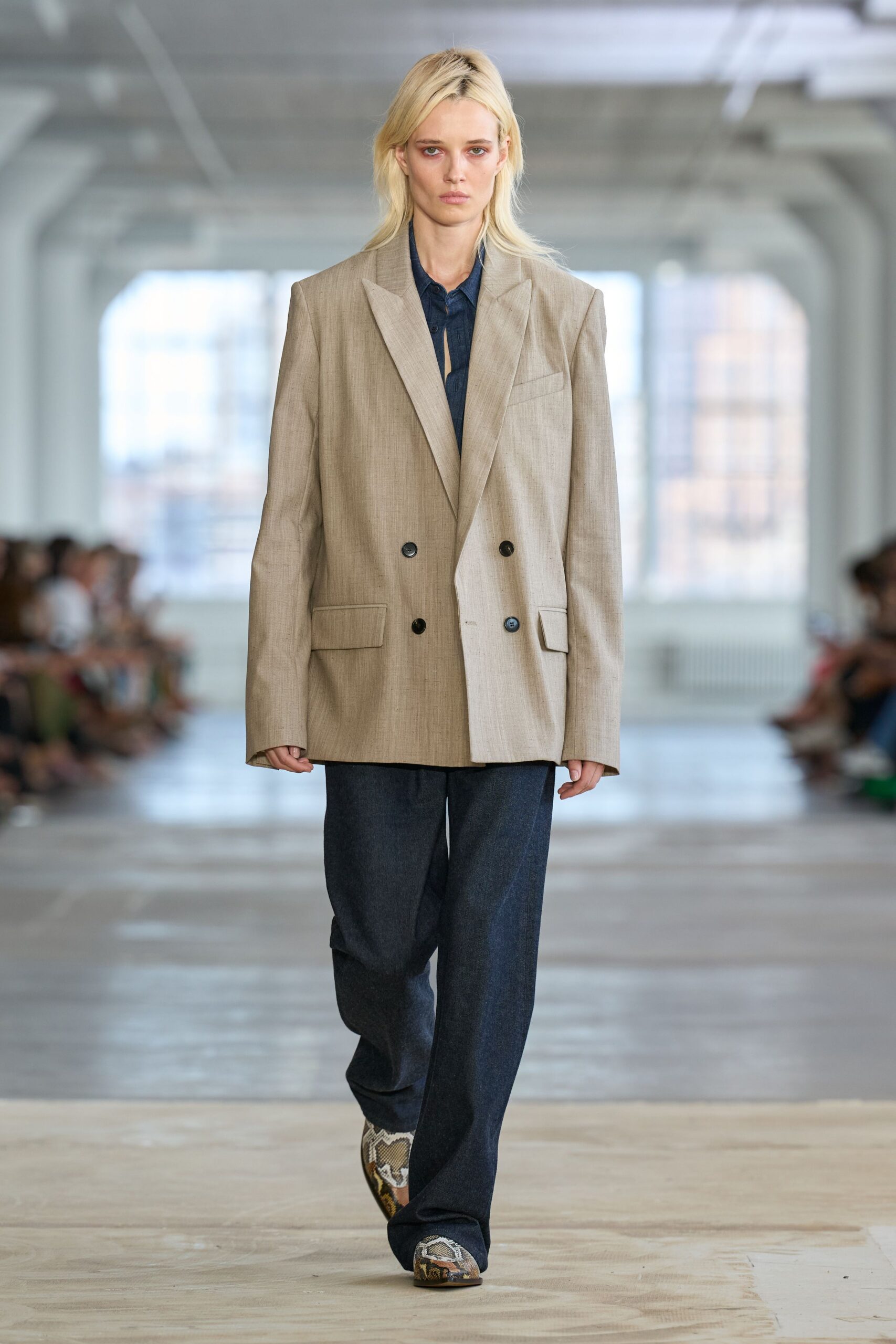 Sand double-breasted blazer with mismatched panels and sharp padded shoulders. Dark indigo shirt, top buttons left open under the lapel break. Charcoal wide-leg trousers with a soft suiting drape. Pointed python boots peeking from the hem. Early train to a pitch meeting, coffee cooling in the paper cup.