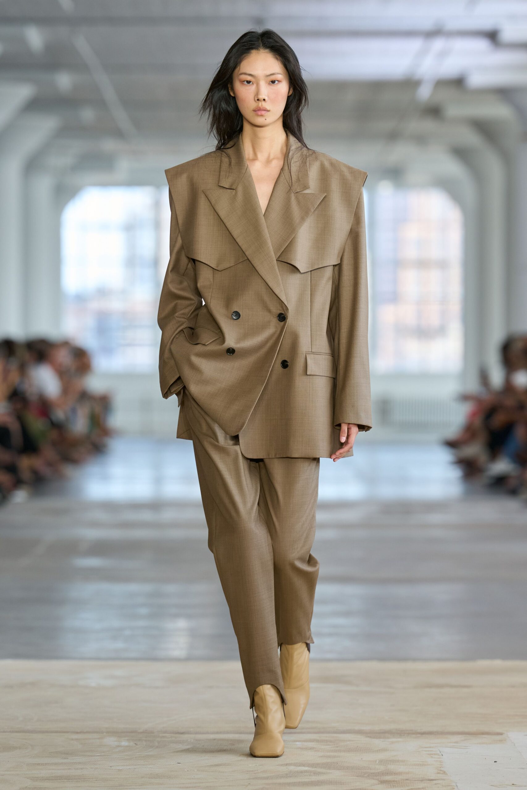 Taupe suiting blazer with exaggerated pagoda shoulders and a folded chest flounce. Matching wool trousers cut loose through the leg, breaking softly at the boot. Cream leather ankle boots with squared toes. Early train to the finance district, coffee cooling in your hand.