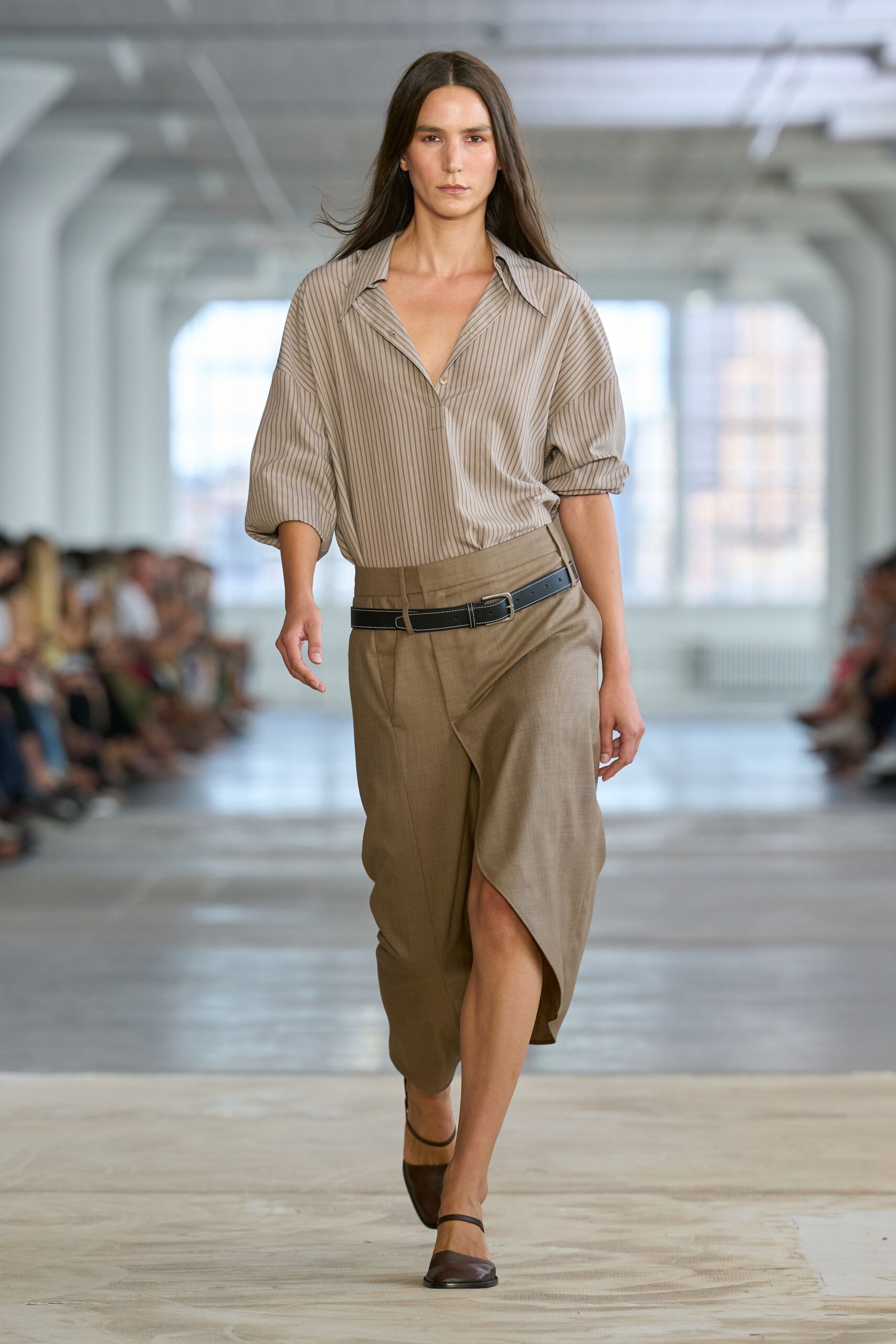 Taupe pinstriped shirt in fluid poplin, sleeves pushed into rounded volume. Mélange tan wrap skirt crossing low at the hip, slit opening mid-stride. Black leather belt with white topstitching and a slack tail. Dark brown ankle-strap flats with squared vamps. Walking into the office on Sunday to water somebody else's plant.