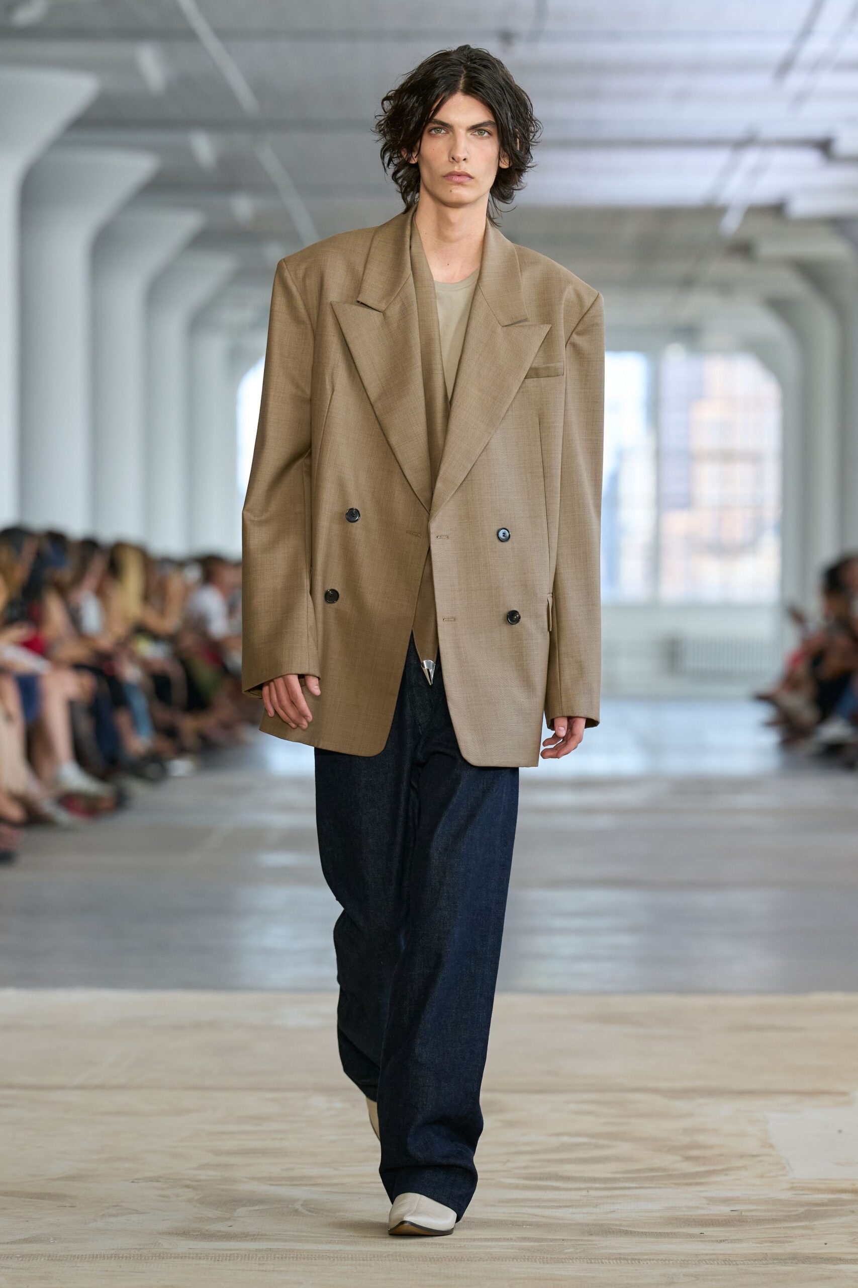 Oversized taupe suiting blazer with peaked lapels and low black buttons. Dusty beige jersey tee, crewneck sitting close under the jacket. Dark indigo wide-leg denim breaking heavy over the shoe. Chalk leather square-toe boots with a sliver of brown sole. Early train to the design office, coffee cooling in a paper cup.