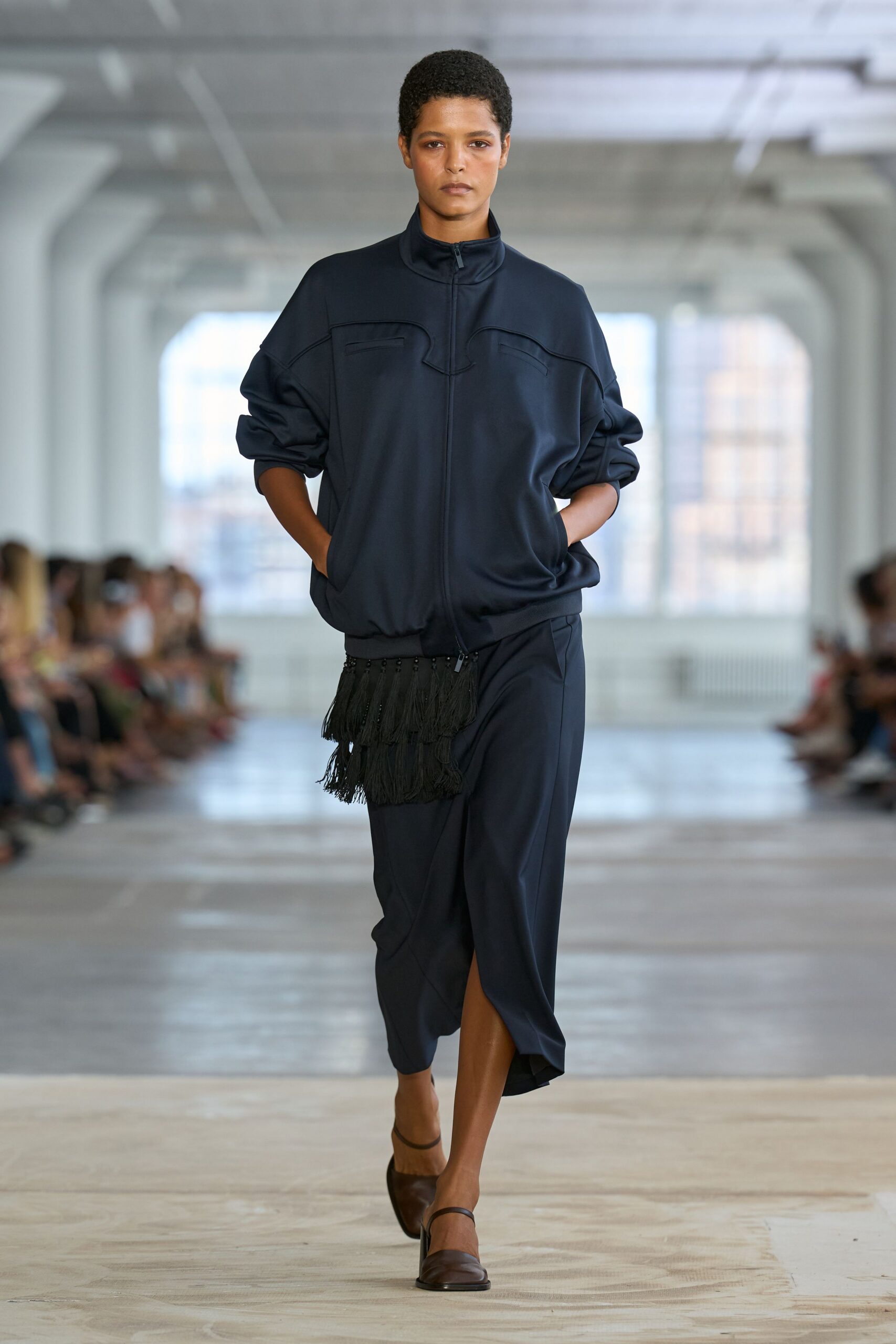 Ink-dark jersey track jacket with a curved yoke and tiny chest welts. Black beaded fringe bag swinging from the hem zip. Matching midi skirt draped close, opening to a front walking slit. Brown leather flats with a narrow instep strap. Crossing a quiet office atrium before anyone else has bad news.