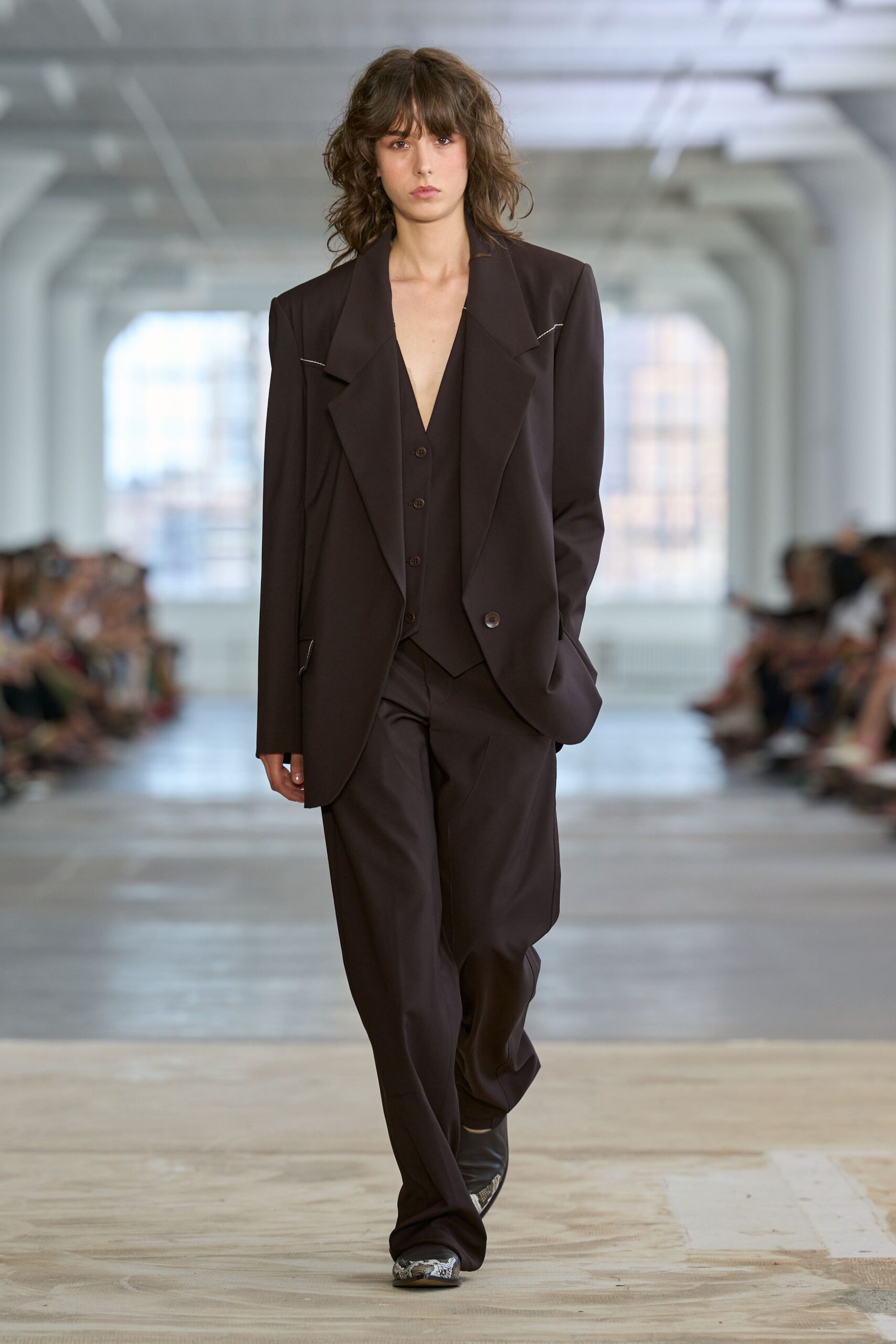 Dark espresso blazer with exaggerated shawl lapels and a stray white basting line. Deep V waistcoat, four-button front cut narrow to a point. Loose tailored trousers breaking softly over the shoe. Pointed python boots flashing from under the hem. Walking the office corridor after everyone else has gone home.
