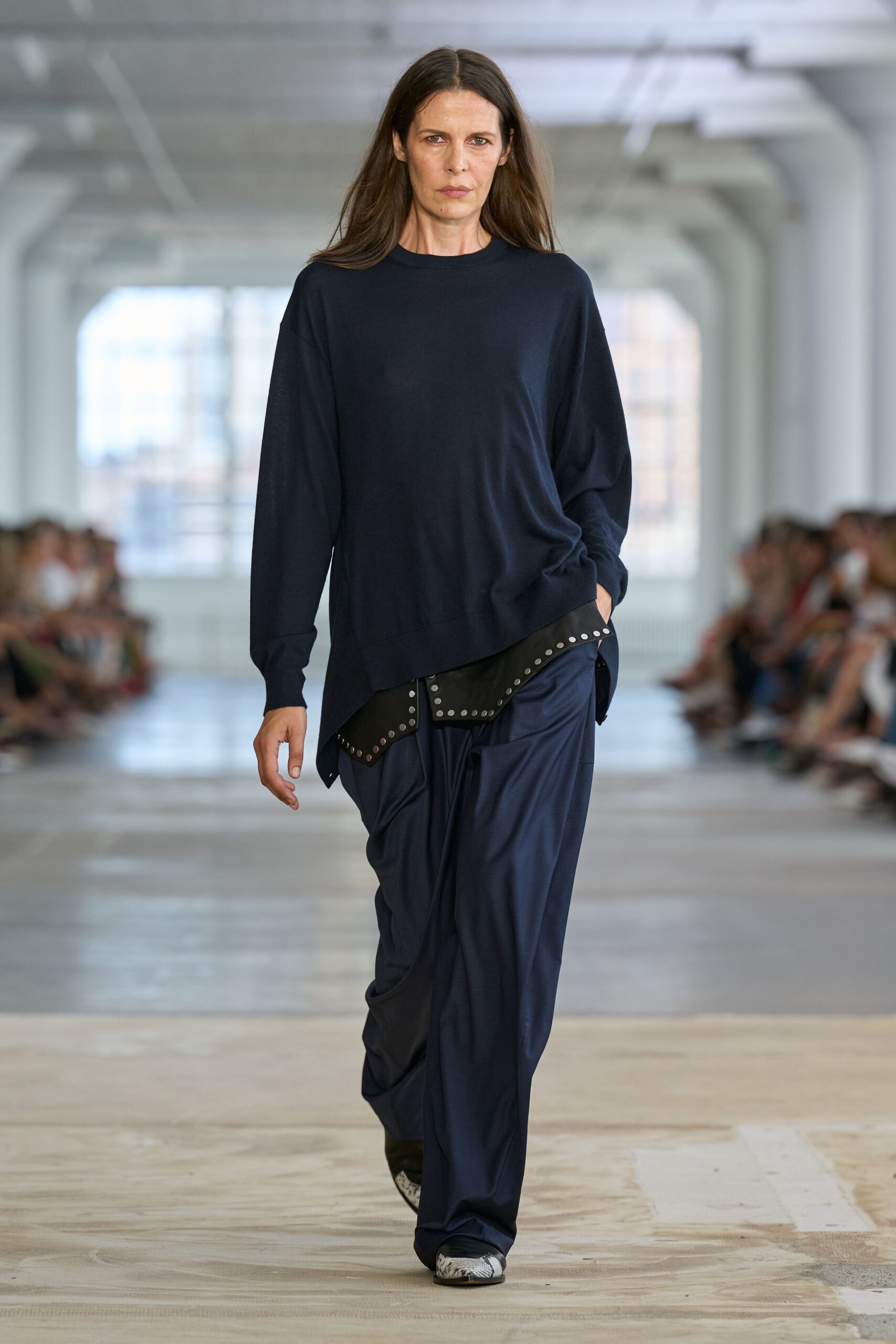 Fine-gauge navy sweater with a skewed hem dropping over one hip. Black leather apron panel rimmed with neat silver studs. Ink trousers cut full and collapsing into deep front folds. Snakeskin square toes peeking from under the break. For crossing a near-empty office on Sunday to grab the charger you forgot.