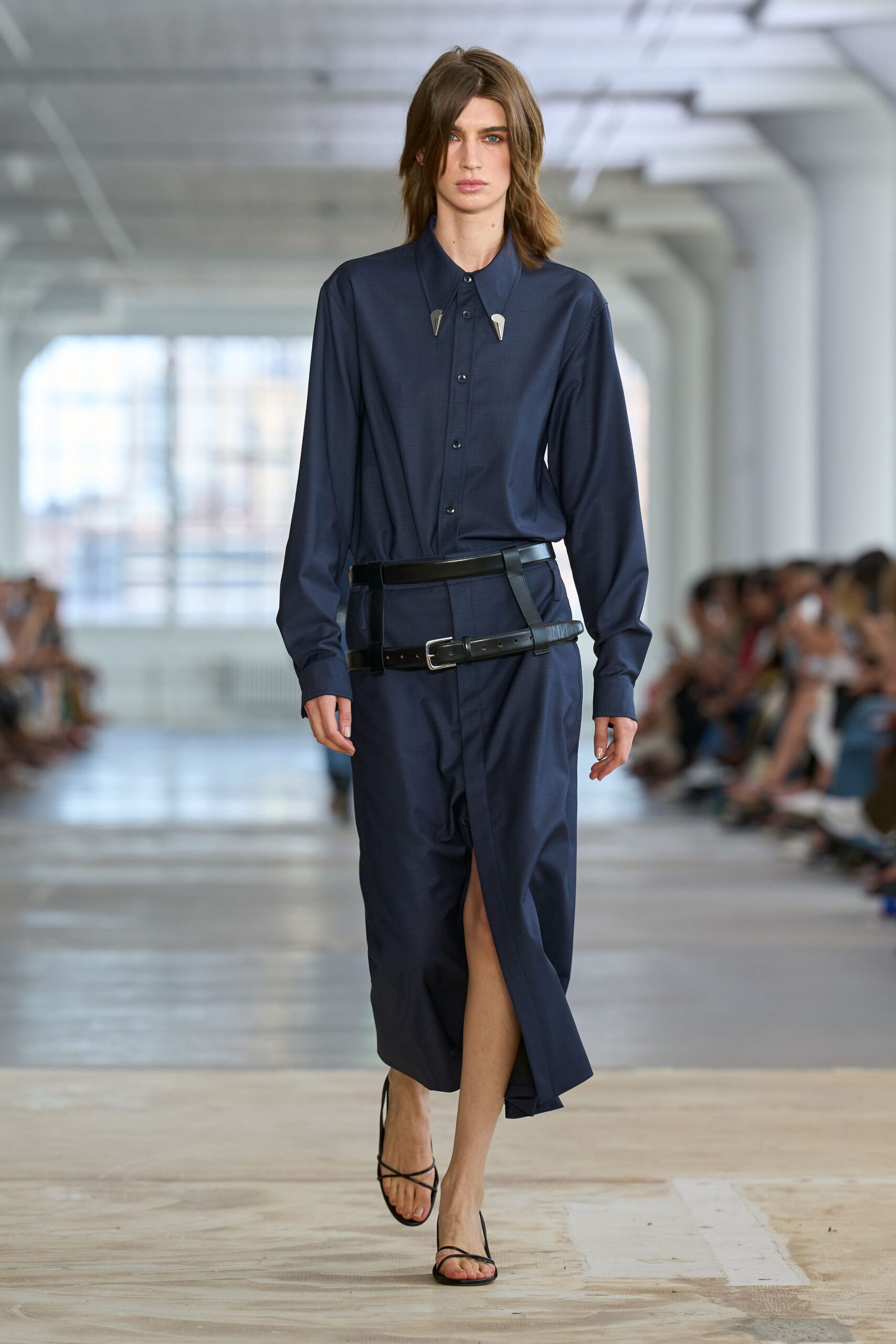 Deep navy shirt in fine microcheck, collar pinned with two pale metal tabs. Black leather belt slung low, extra strap dropping past the buckle. Matching wrap skirt opening to a narrow front slit below the knee. Bare black thong sandals with thin ankle straps. For crossing a near-empty office tower lobby after everyone else left.