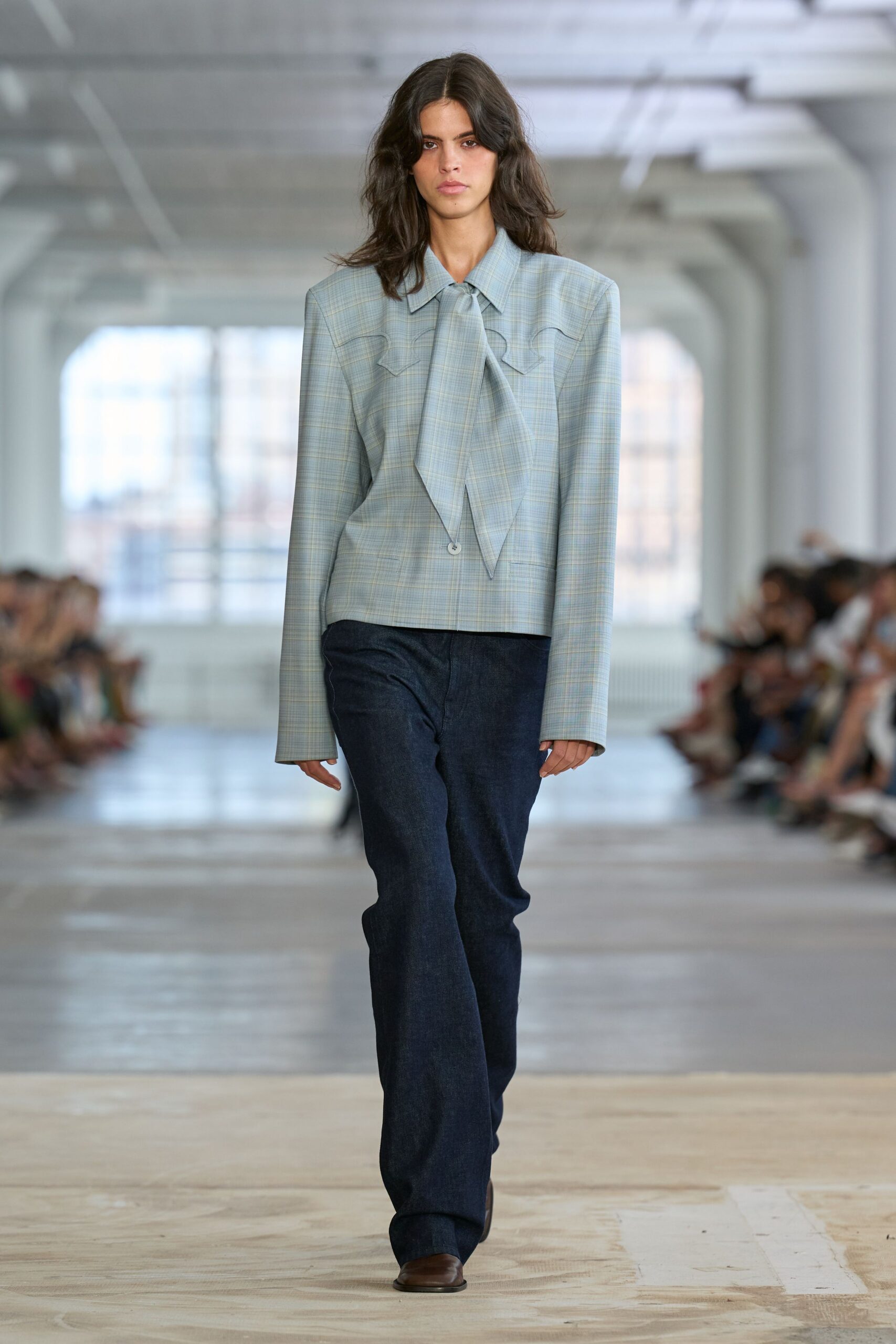Powder-blue plaid blouse with a long scarf tie and curved yoke seams. Dark indigo jeans cut straight and loose over the instep. Square brown leather toes peeking from beneath the hem. Early train to the county courthouse, paperwork folded in a tote.
