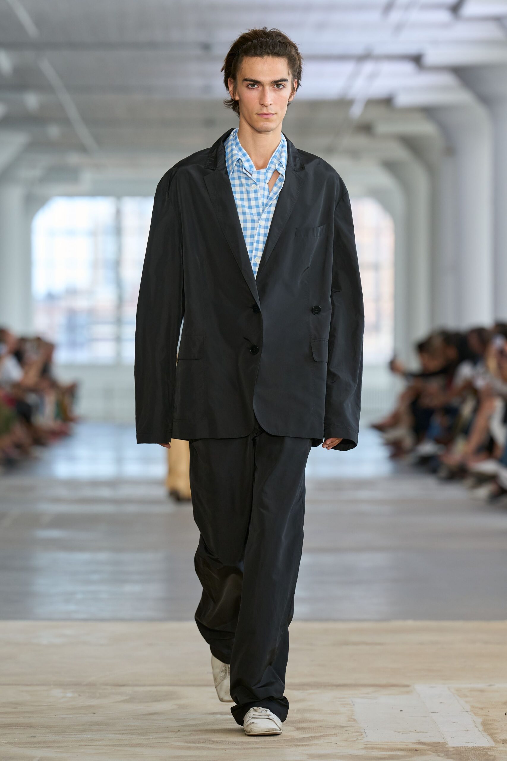 Black nylon blazer cut oversized, sleeves falling past the hands. Sky-blue gingham shirt, one side of the collar slipping open. Loose black trousers in the same faintly shiny nylon. White sneakers, creased and greyed at the toes. First day at a borrowed office, still carrying your real life in a tote.