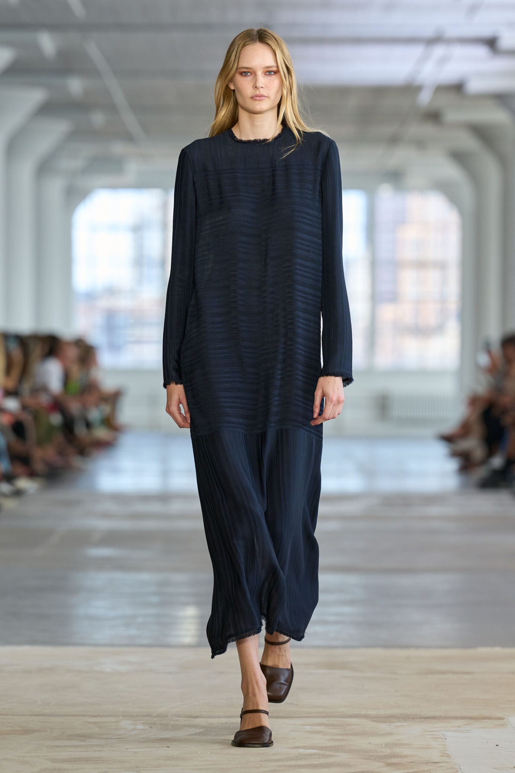 Ink-black crinkled tunic, horizontal puckering across the body, raw fringed edges. Matching pleated trousers falling loose to a softly frayed cropped hem. Brown square-toe slingback flats with slim ankle straps. Walking the echoing corridor before offices fill and the lights fully wake up.