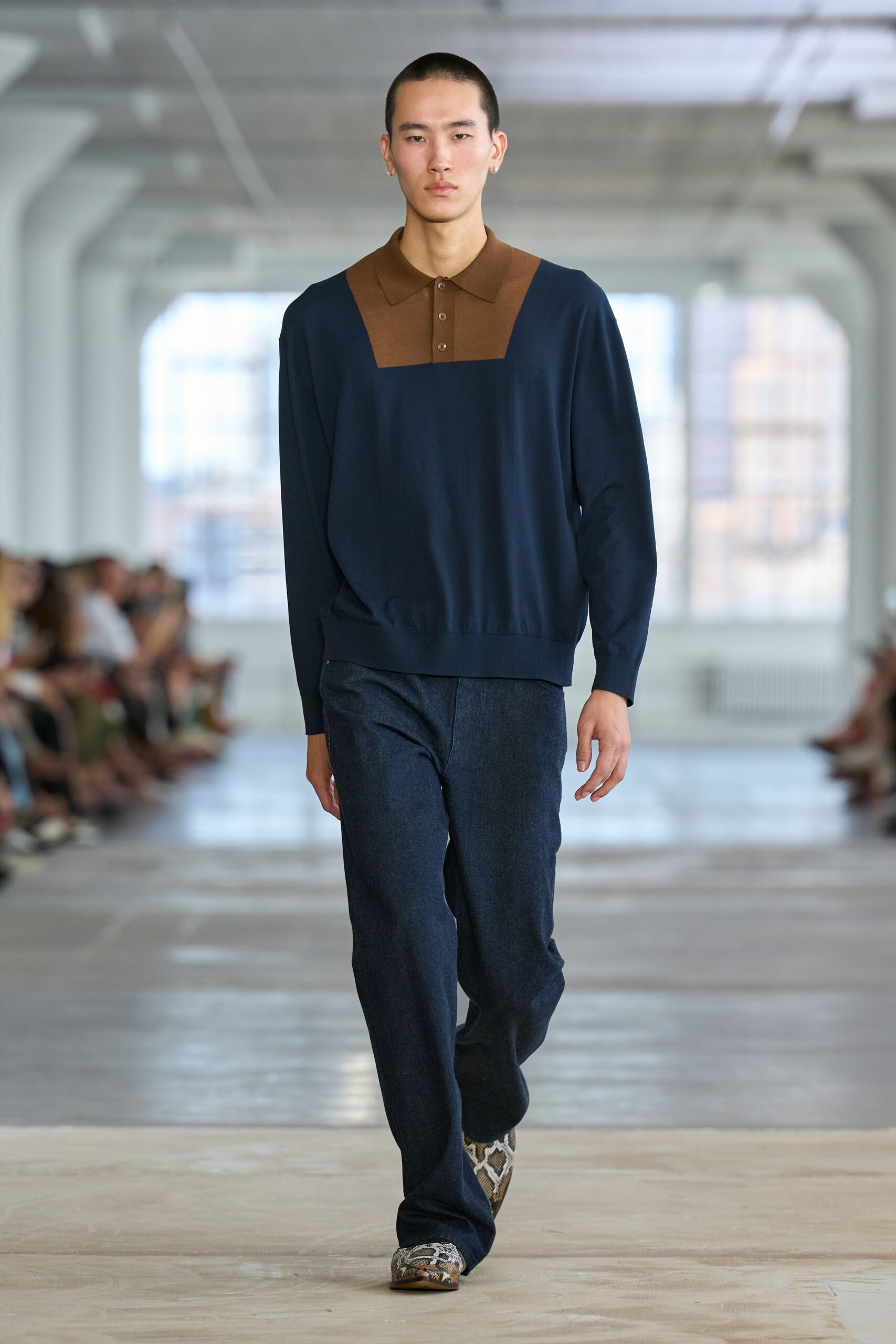 Navy fine-gauge knit polo with a chestnut bib panel and three-button placket. Dark indigo trousers in a softly flecked weave, falling almost straight. Python boots with squared toes peeking from the hem. Office tower Sunday, nobody else on the floor but the cleaners.